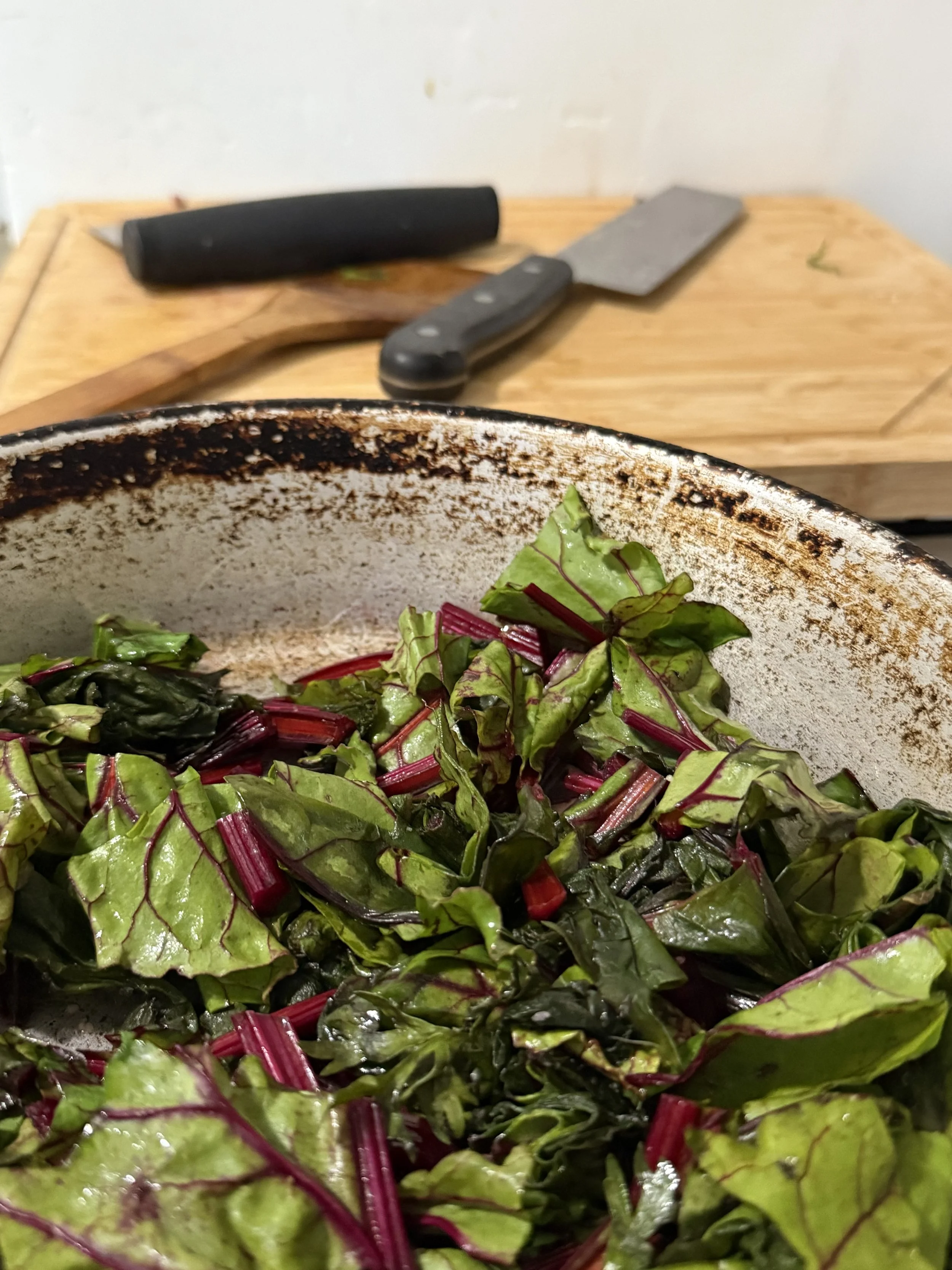 Leafy greens cooking in a pan with a cutting board and kitchen knives behind