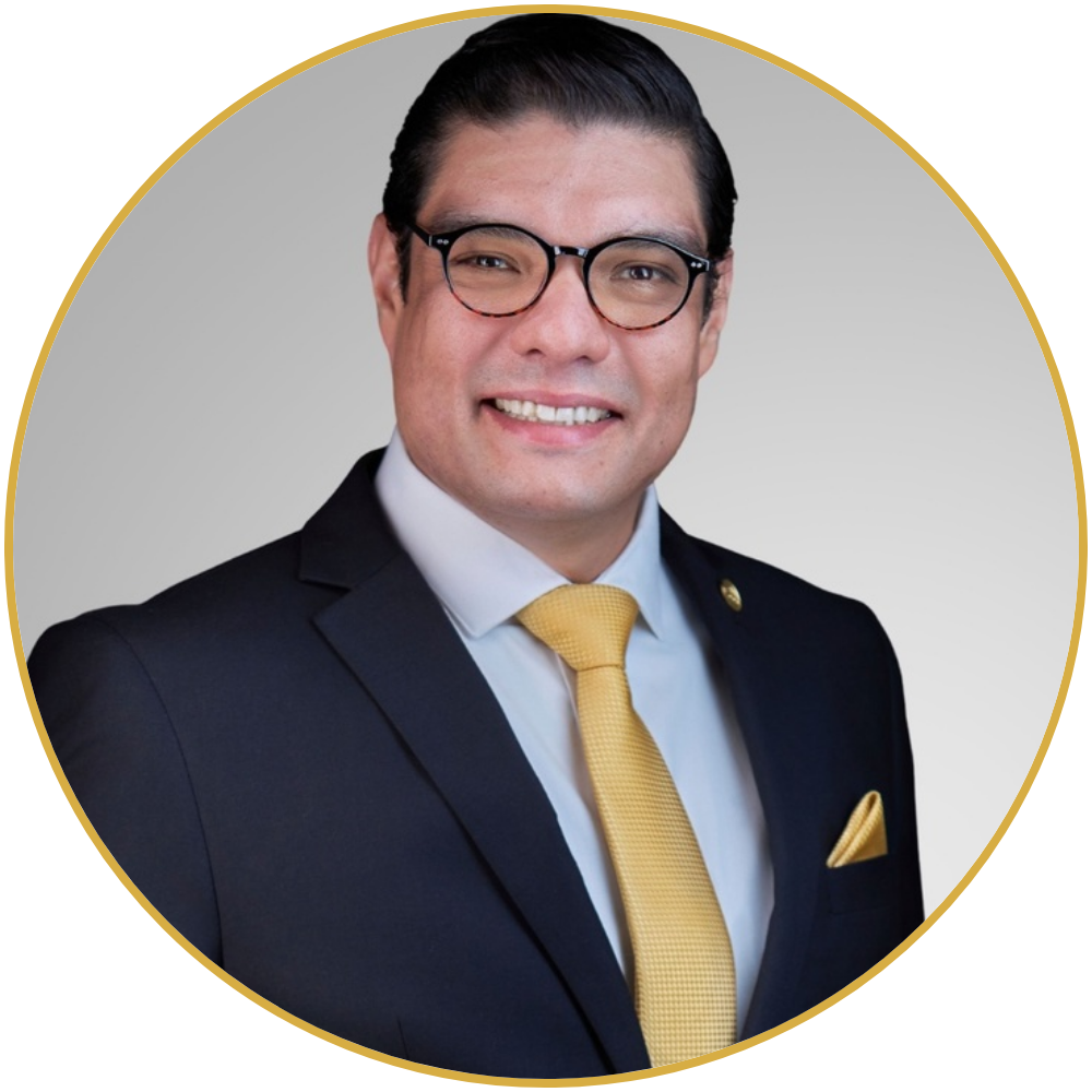 A professional man smiling, wearing glasses, a dark suit, a white shirt, and a yellow tie and pocket square, against a plain gray background.