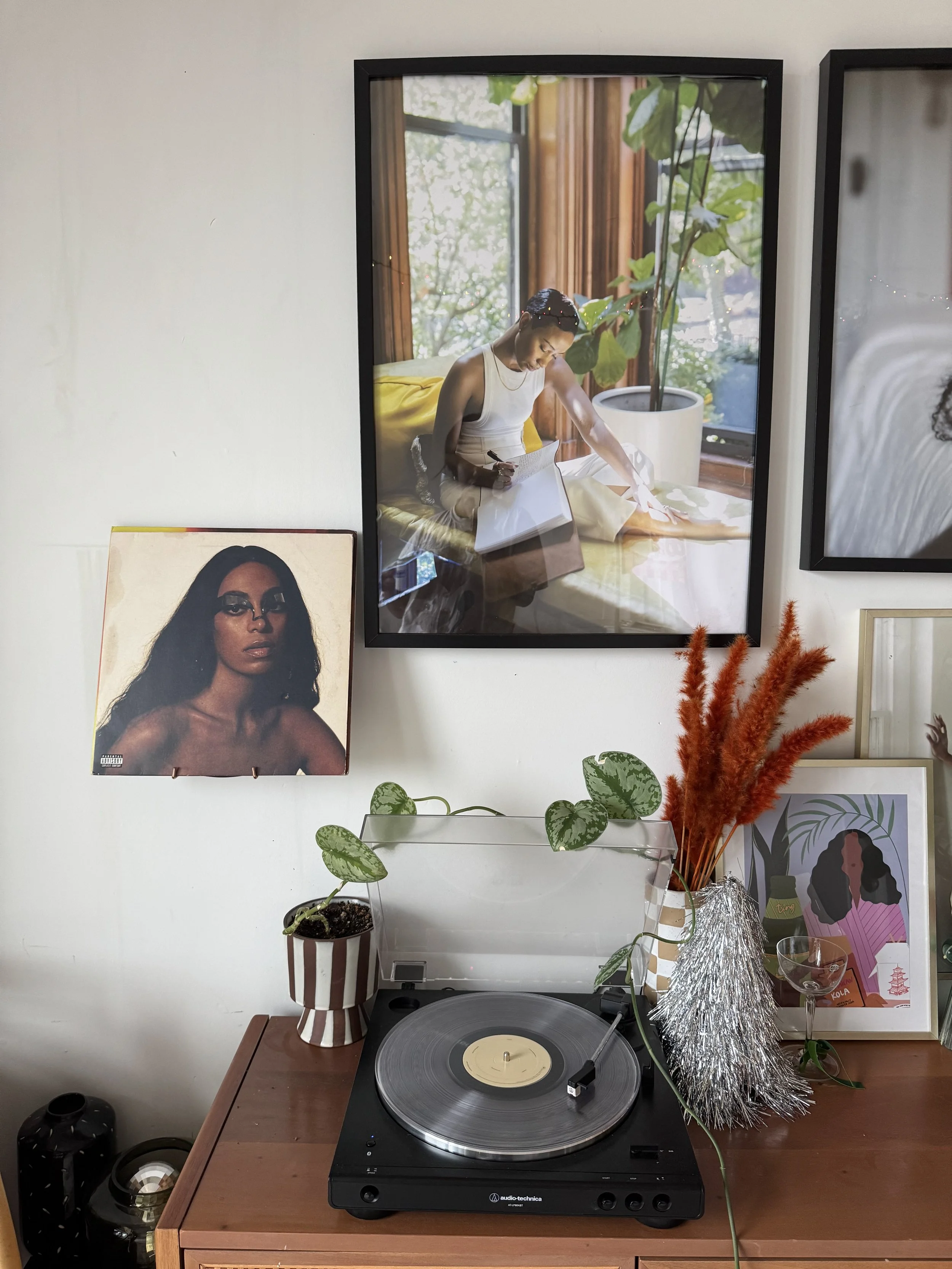 A wooden table with a turntable and a potted plant, alongside decorative orange feathers and a framed colorful portrait of a woman with dark hair. Above the table, two framed photographs are hung on a white wall. One photograph shows a woman sitting 