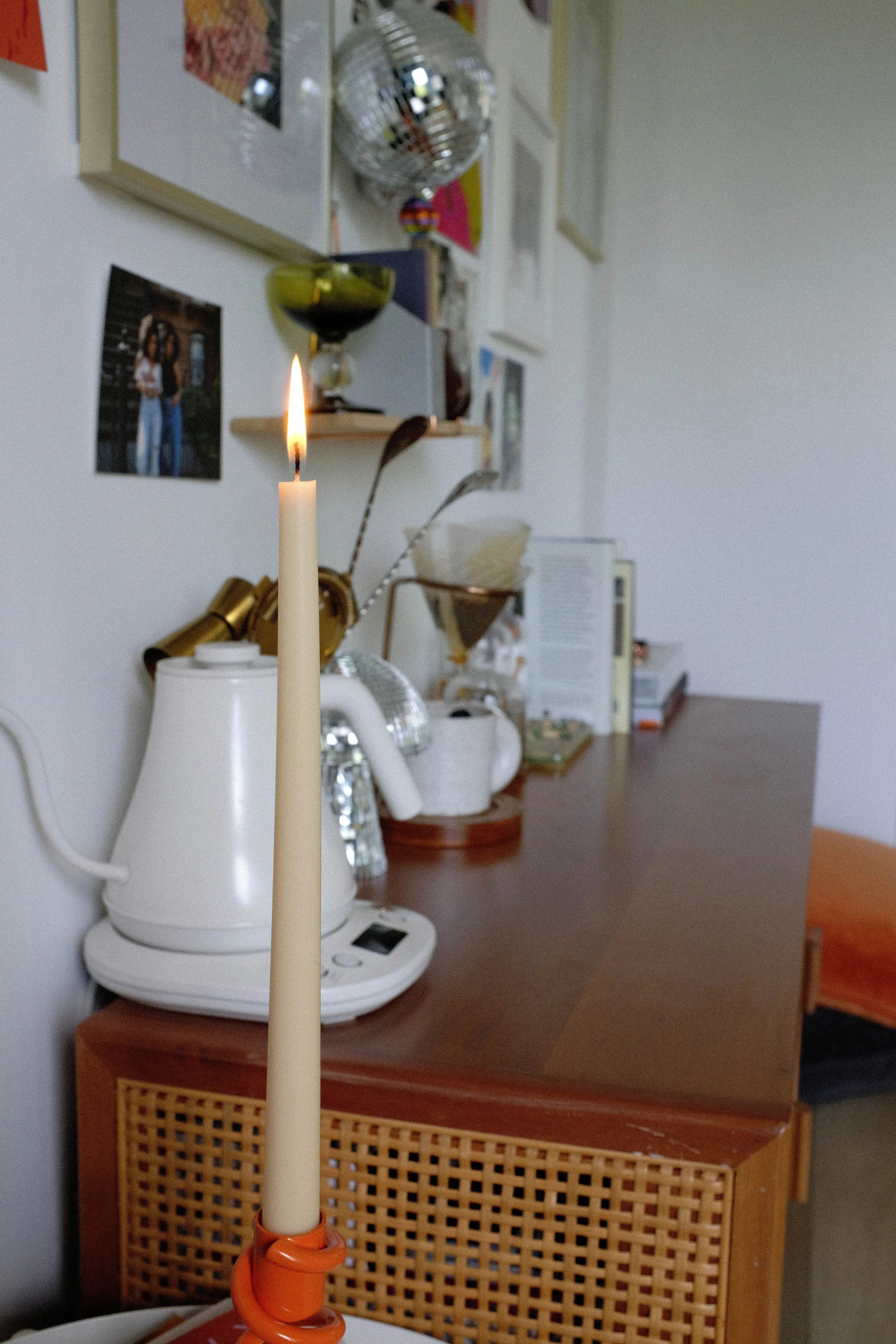 A lit candle on a table in a room with a wooden sideboard, appliances, and various decorative items on the wall including photographs and disco ball.