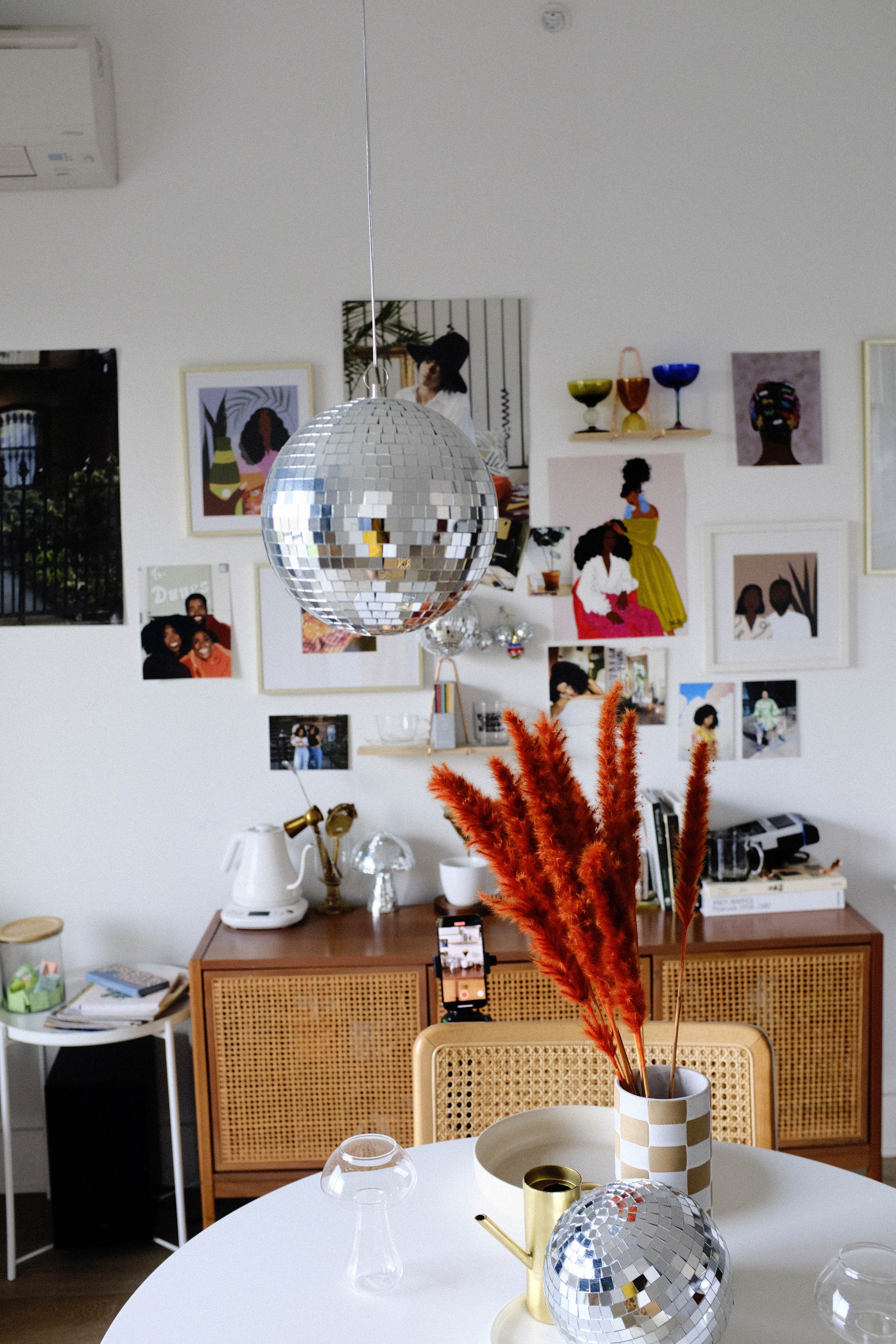 A room with a white wall decorated with various art prints and photos, a wooden cabinet with a white kettle, books, and decorative items, and a table with a checkered vase holding orange-red pampas grass, a gold watering can, and a disco ball.