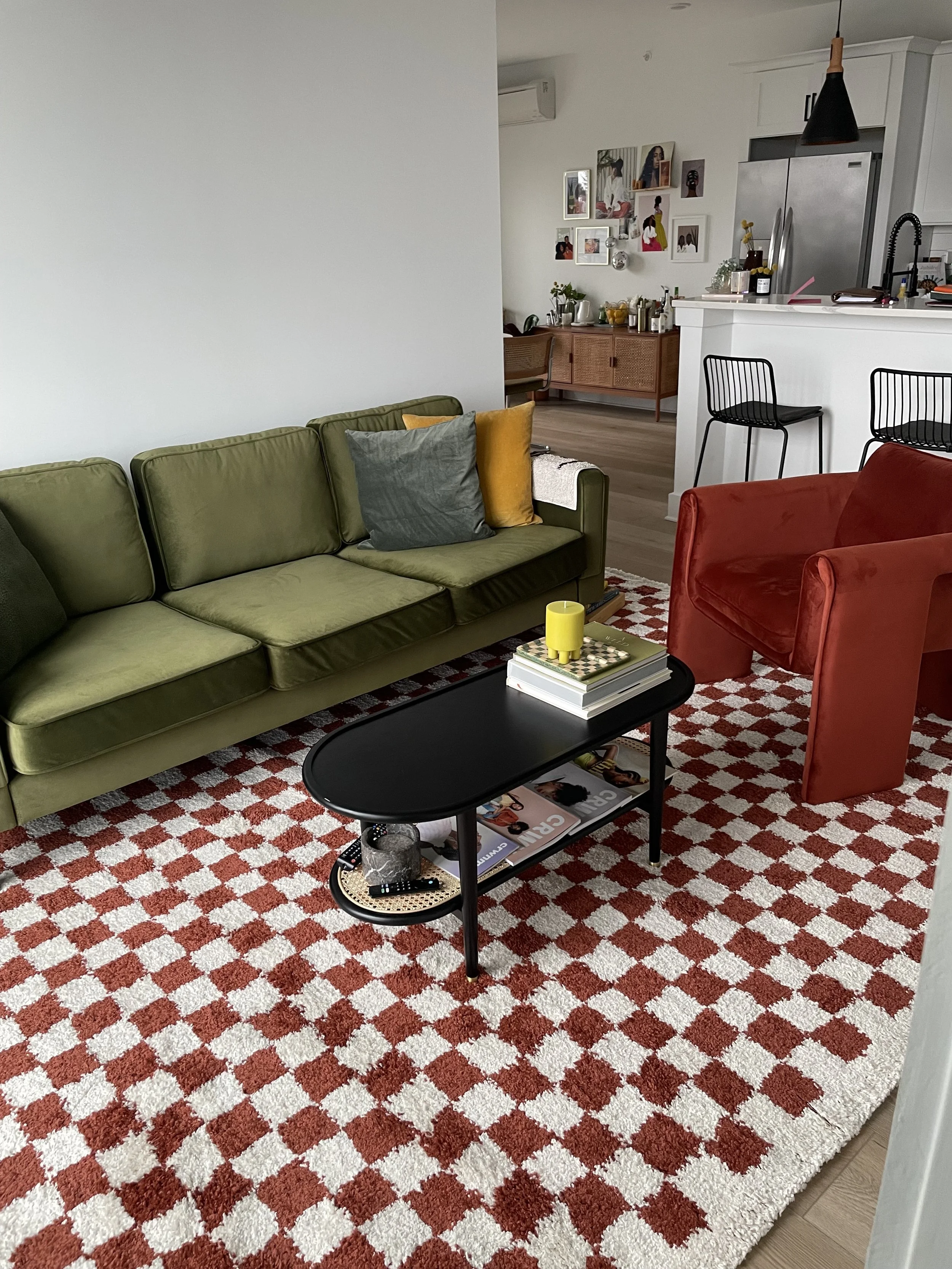 Living room with a green sofa, red armchair, black coffee table with magazines and a yellow candle, red and white checkered rug, and an open kitchen in the background.