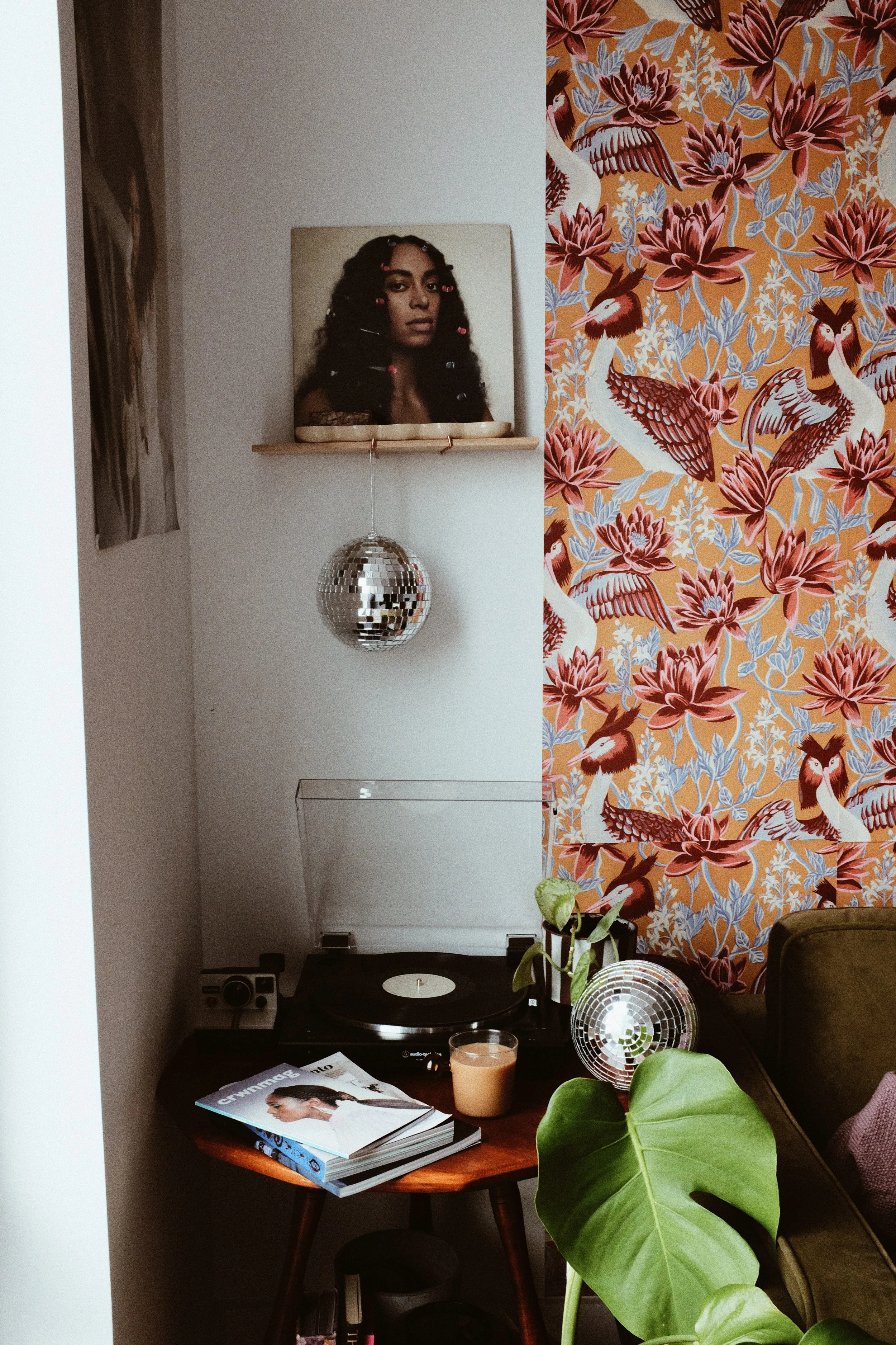 A cozy corner with a turntable on a wooden table, a magazine, a candle, and a disco ball. A large green plant with heart-shaped leaves is in front of the table. The background features a white wall, a colorful floral and bird patterned wallpaper, a p
