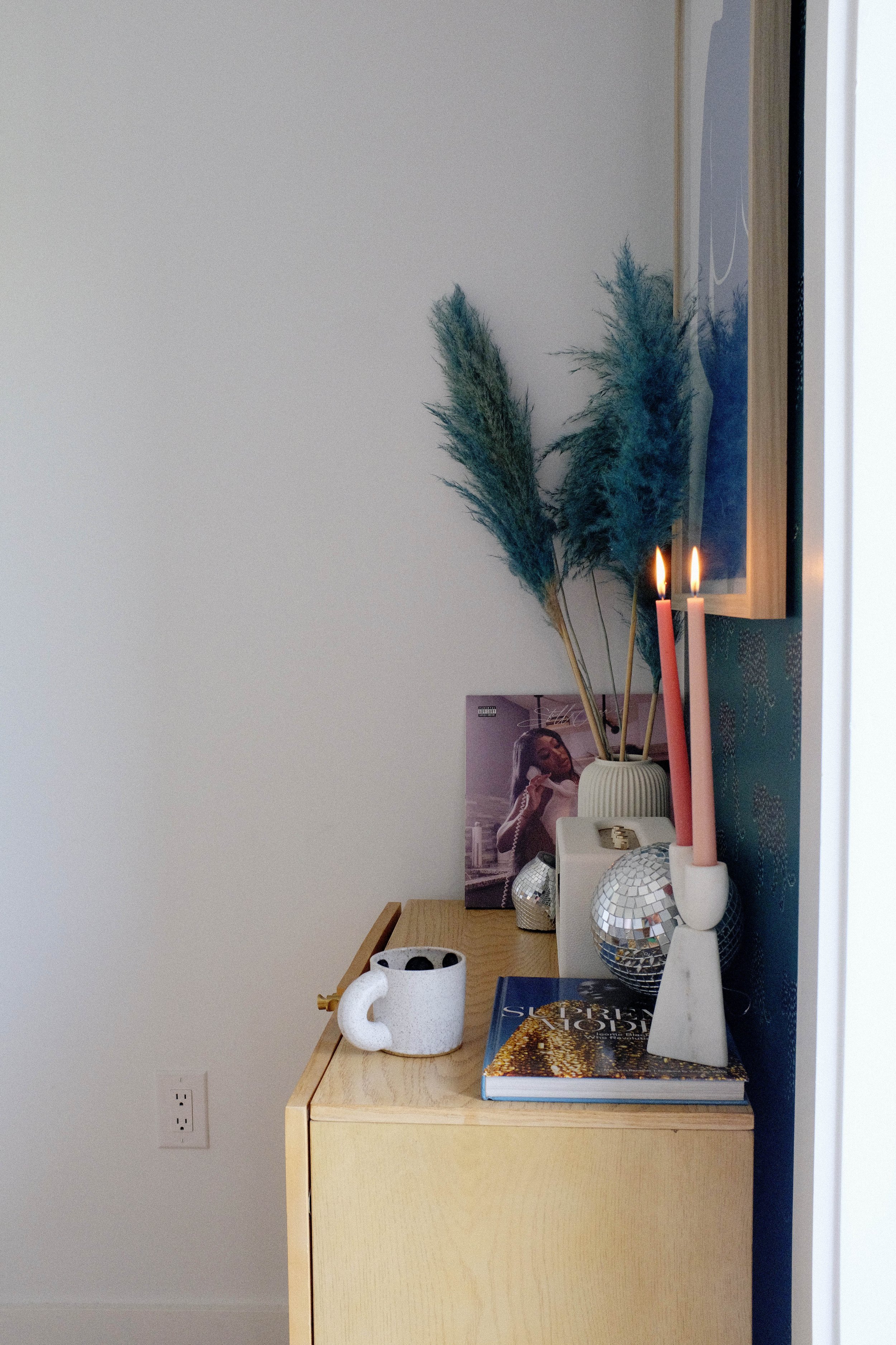 Decorative items on a wooden dresser, including a white cup, bookshelf, candle holders with pink candles, and a artwork reflected in a mirror with green stems of pampas grass in a white vase.