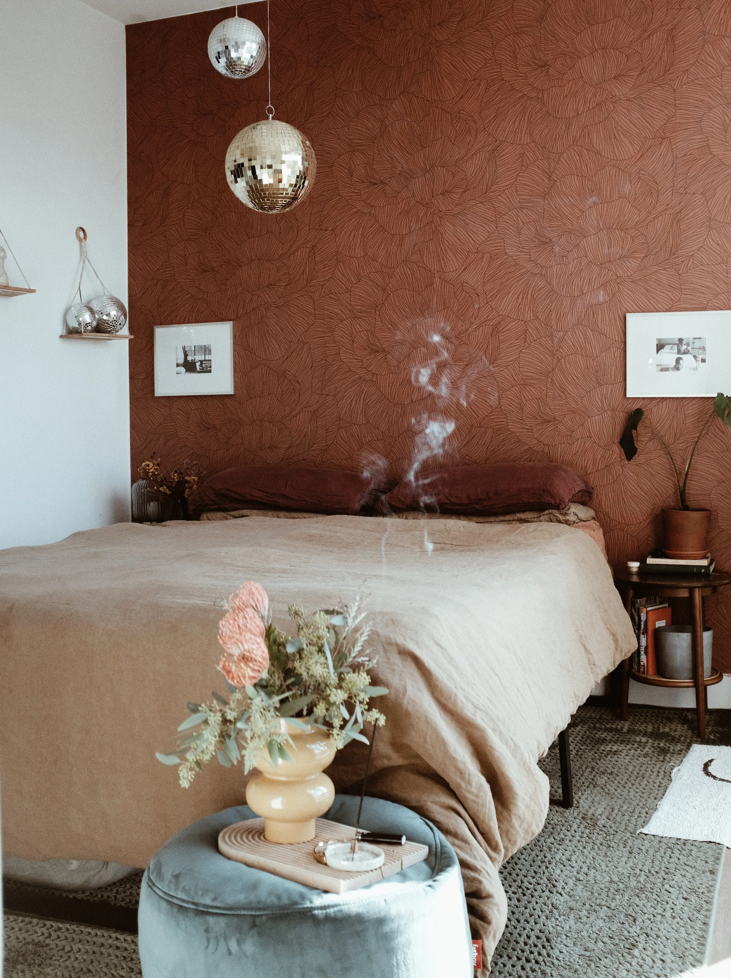A cozy bedroom with a beige bed, rust-colored patterned accent wall, hanging disco balls, framed photos, a potted plant, and a small side table with books and decor. There is a vase with flowers on a small round table in the foreground.