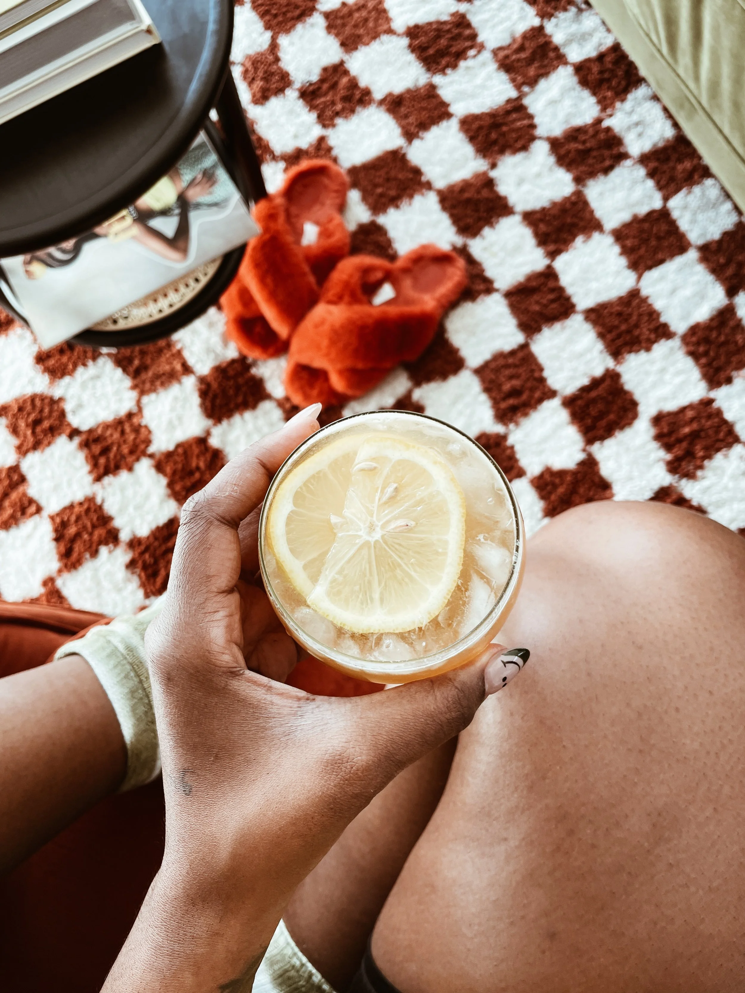 A person sitting on a chair or sofa holding a glass of lemonade with lemon slices. On the floor, there are a pair of fuzzy orange slippers. A checkered carpet and a small table with a magazine are also visible.