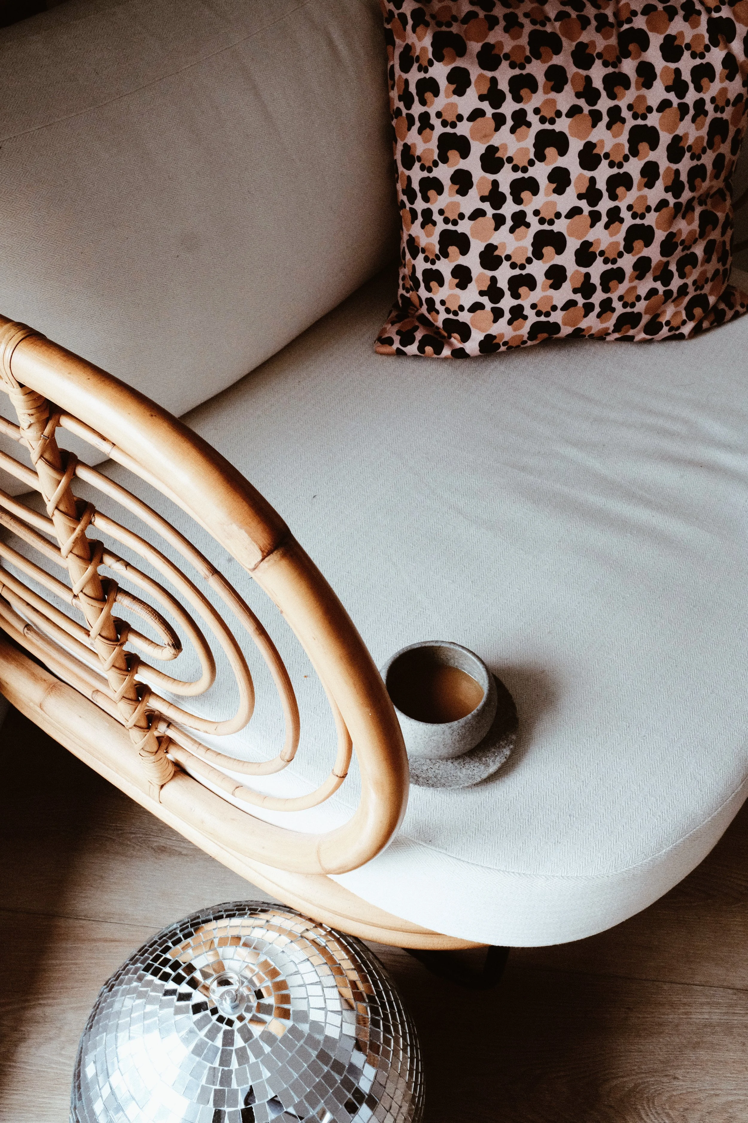 A beige sofa with a rattan armrest, a leopard print pillow, a small round coffee table with a cup of coffee, and a disco ball on the floor.