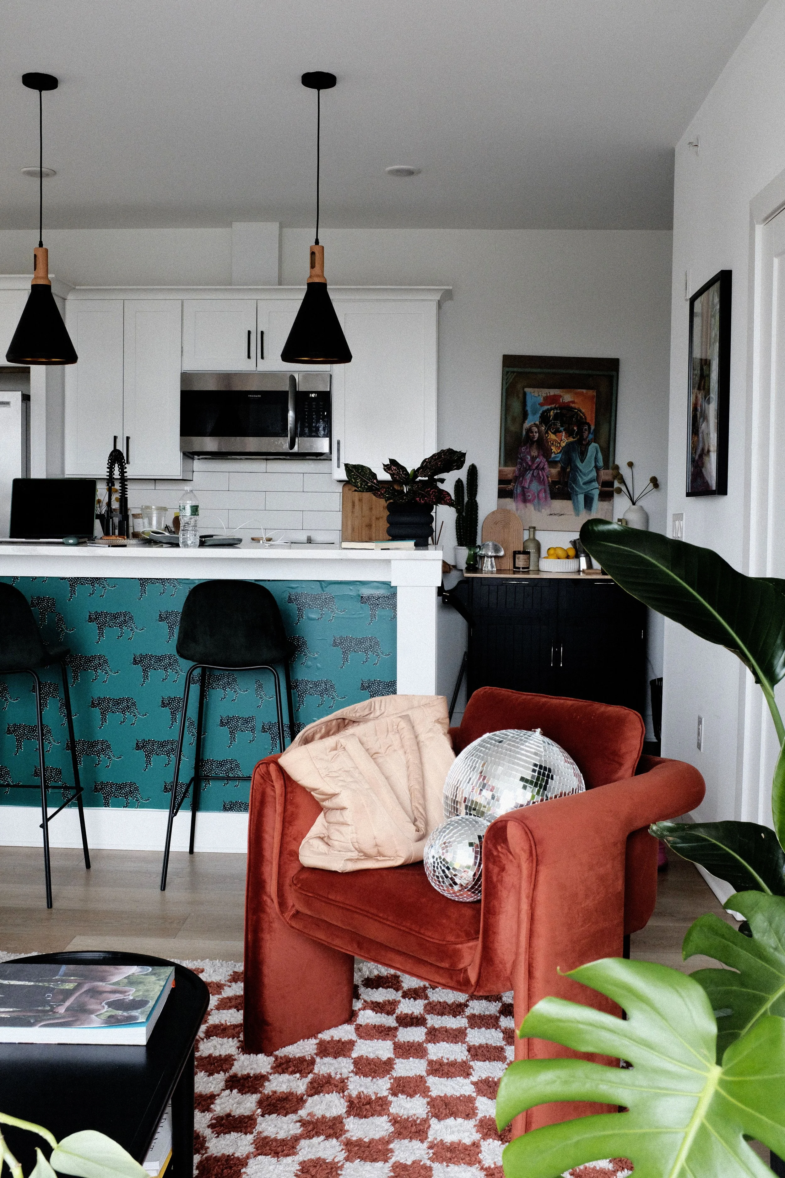 Living room with a red armchair, balloons, green plants, and a patterned rug in front of an open kitchen with white cabinets and black chairs.