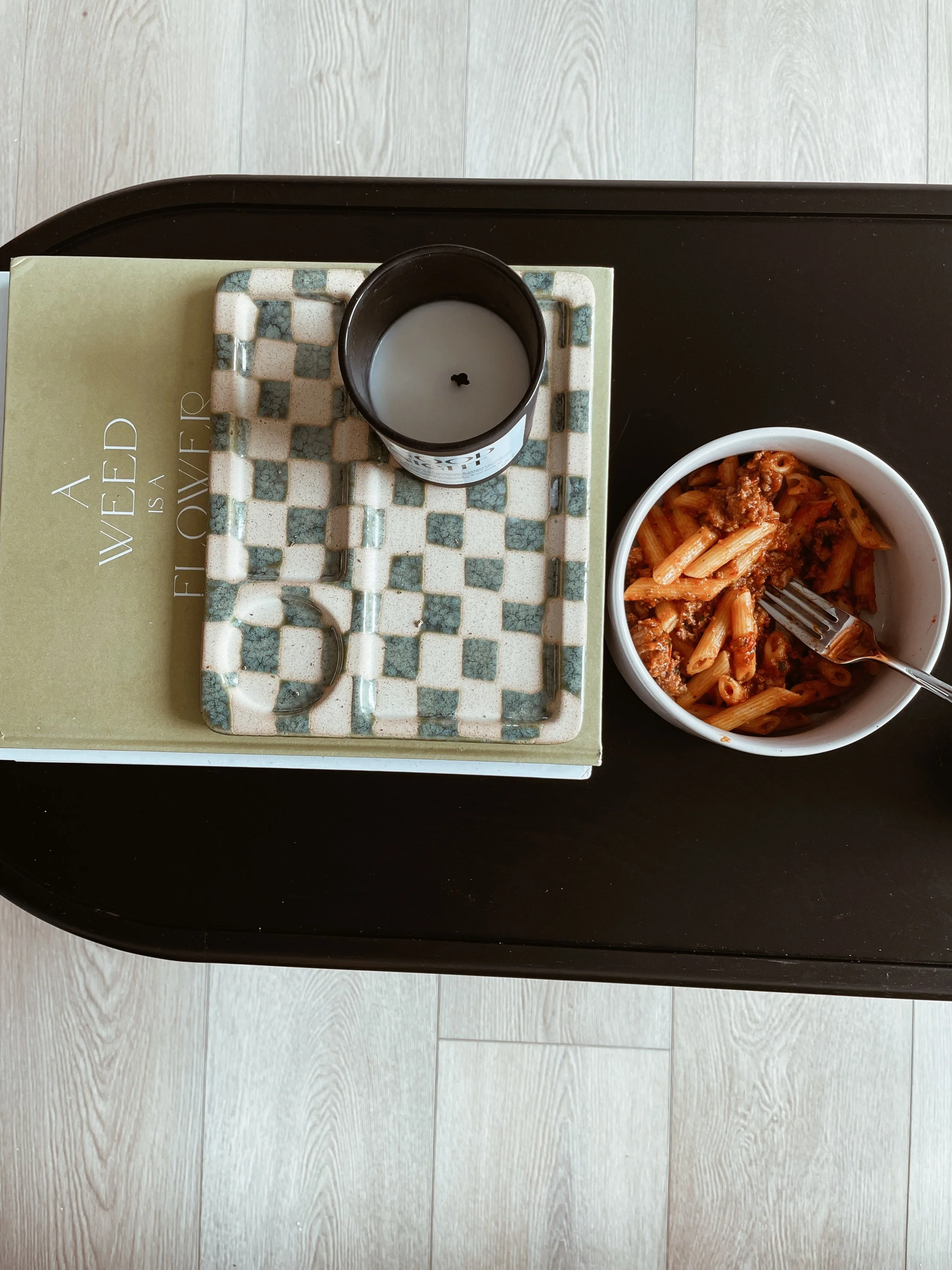 A tray with a candle, a book titled 'A WED is a FI OVE', and a bowl of pasta on a dark table, viewed from above.