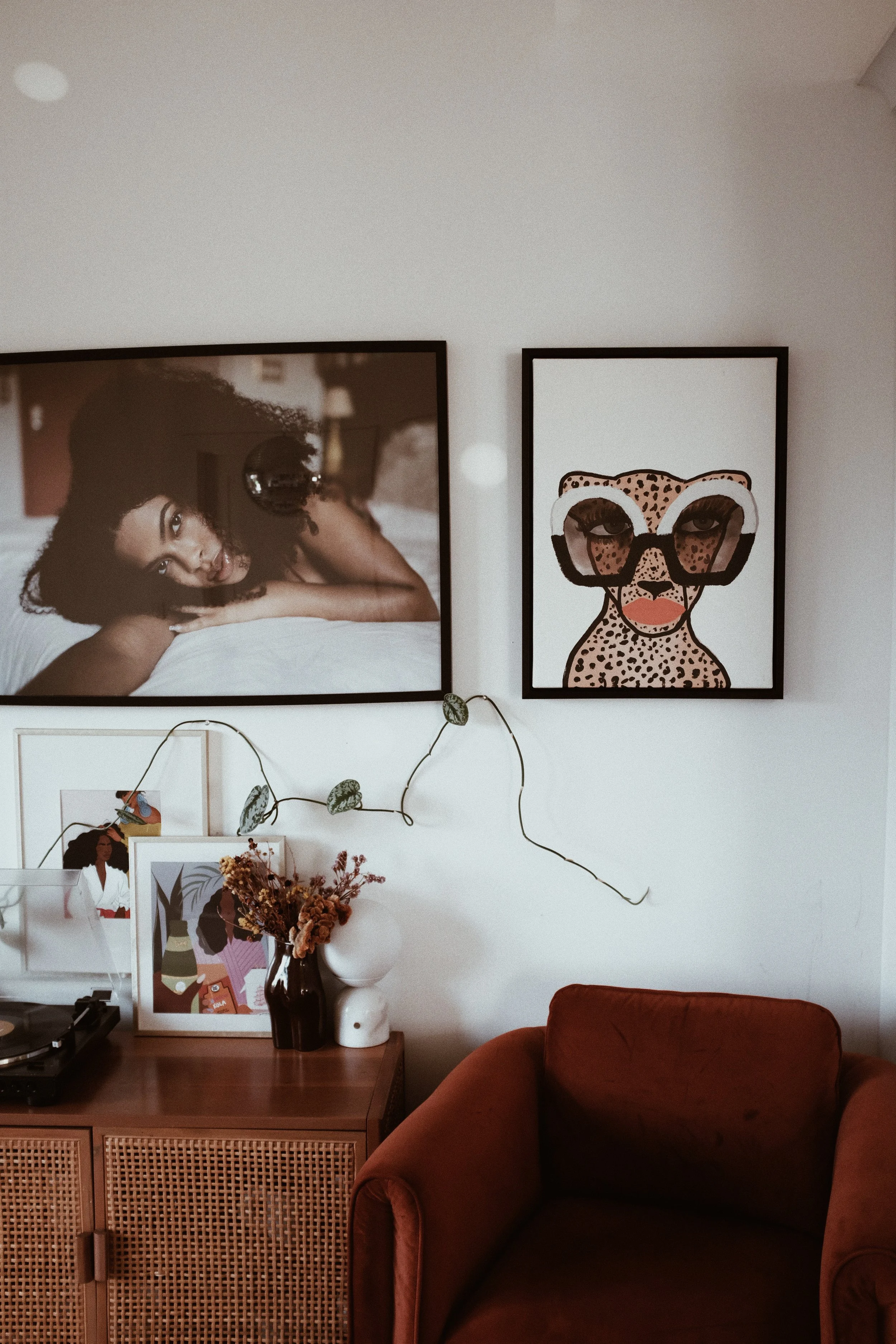 Living room with two framed art prints on the wall: one of a woman lying on her side, and the other of a cheetah wearing glasses. Below the art, there is a rattan cabinet with vases, framed pictures, dried flowers, and a turntable. A brown armchair i