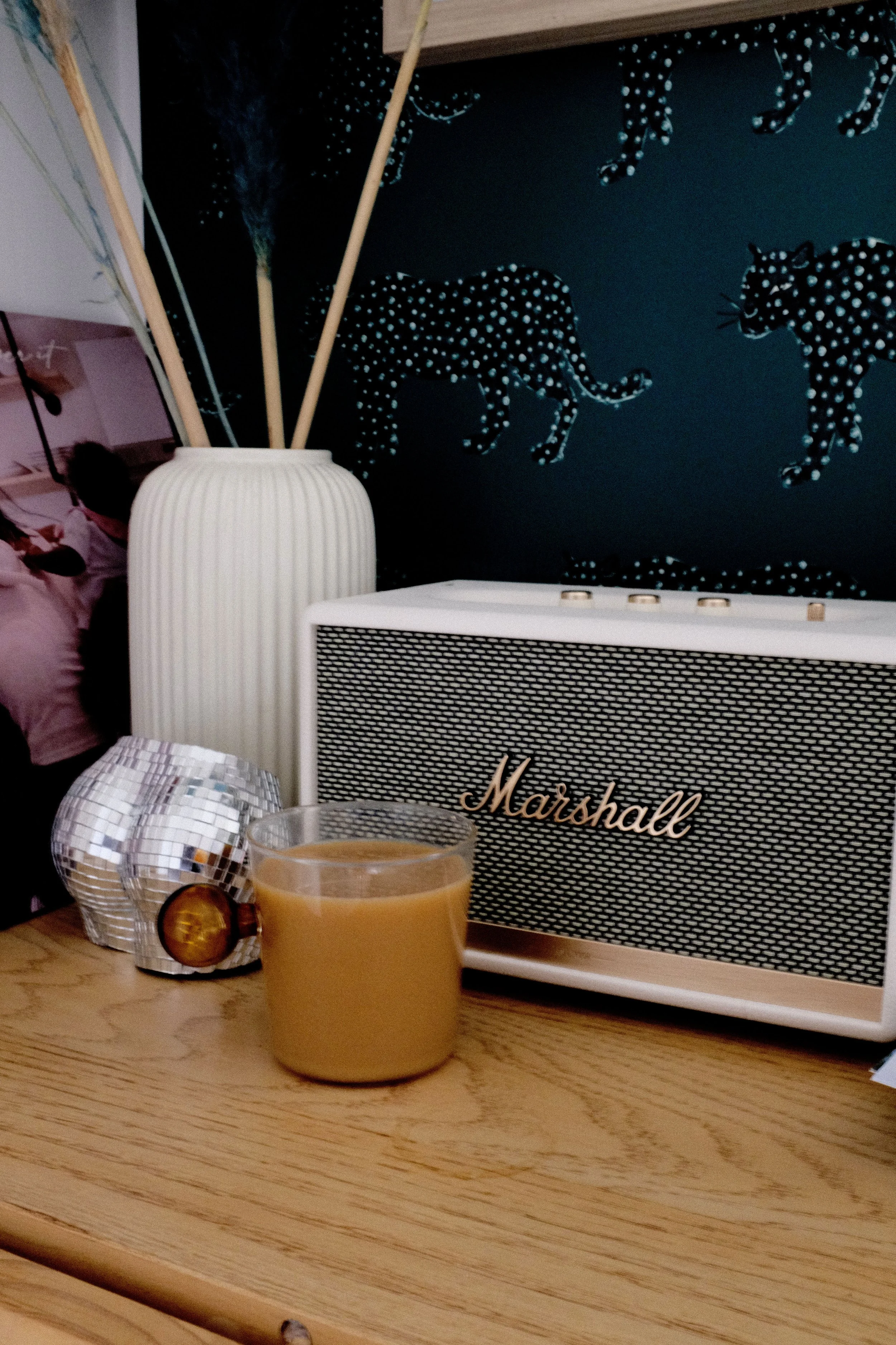 A wooden table with a glass of coffee, a decorative disco ball, a white Marshall speaker, a silver vase with sticks, and a blue wall with animal-shaped neon light art.
