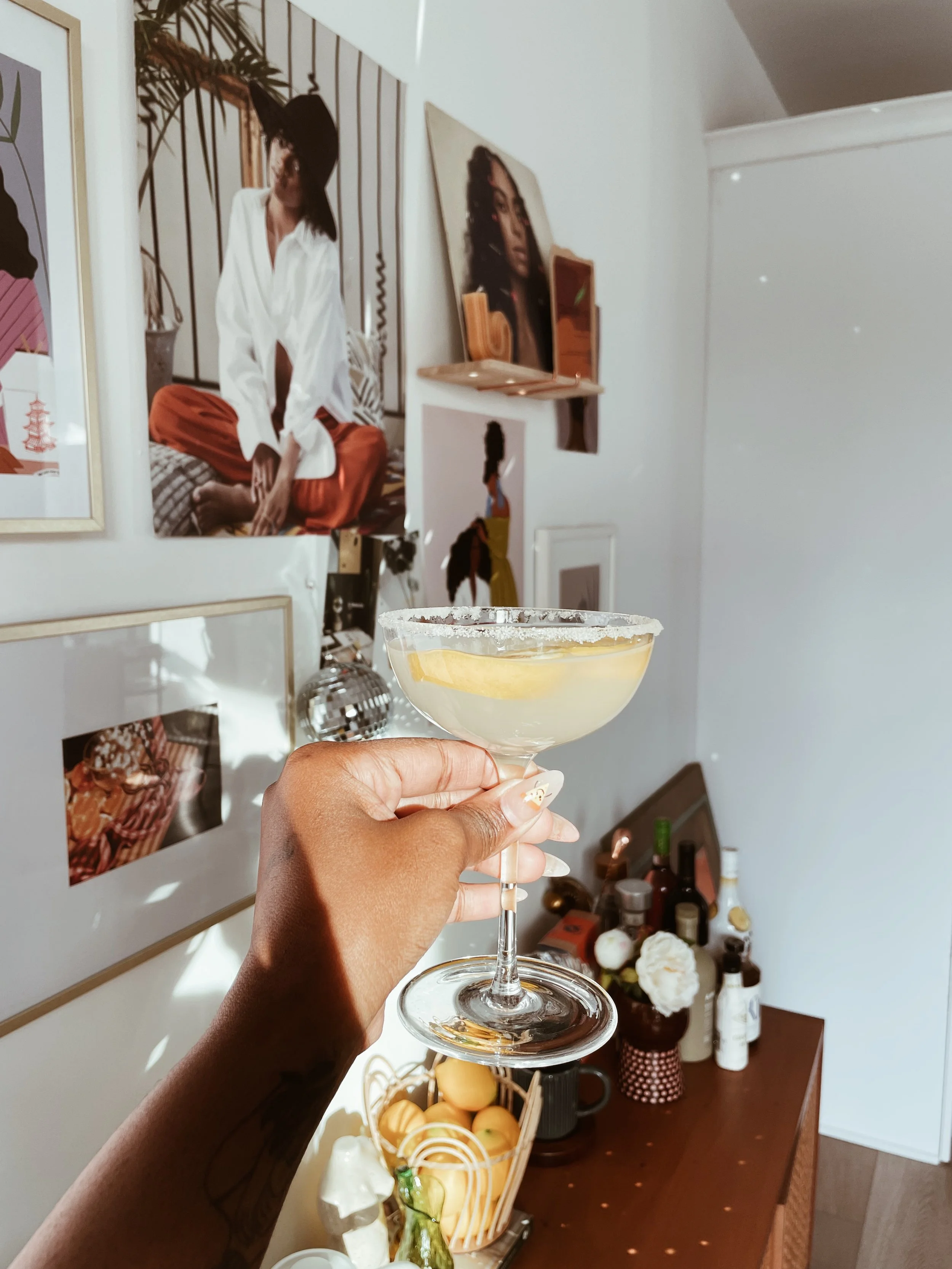 A hand holding a cocktail glass with a lemon slice inside, in front of a decorated wall with framed photos and artwork.