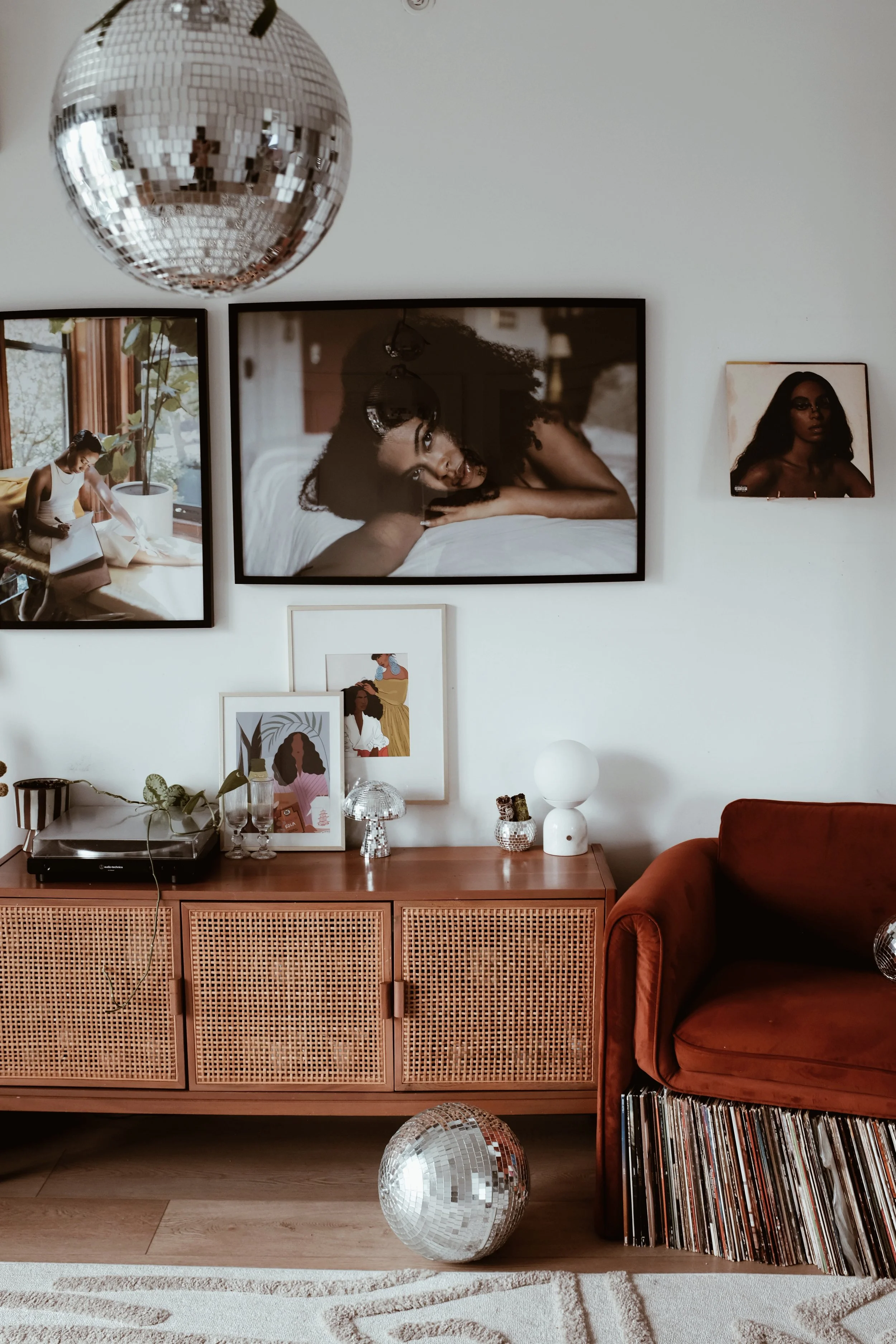 A living room interior featuring a wooden sideboard, a red velvet sofa, and various wall art including photos and illustrations of women. A disco ball is hanging from the ceiling, with another disco ball on the floor. The sideboard has decorative ite