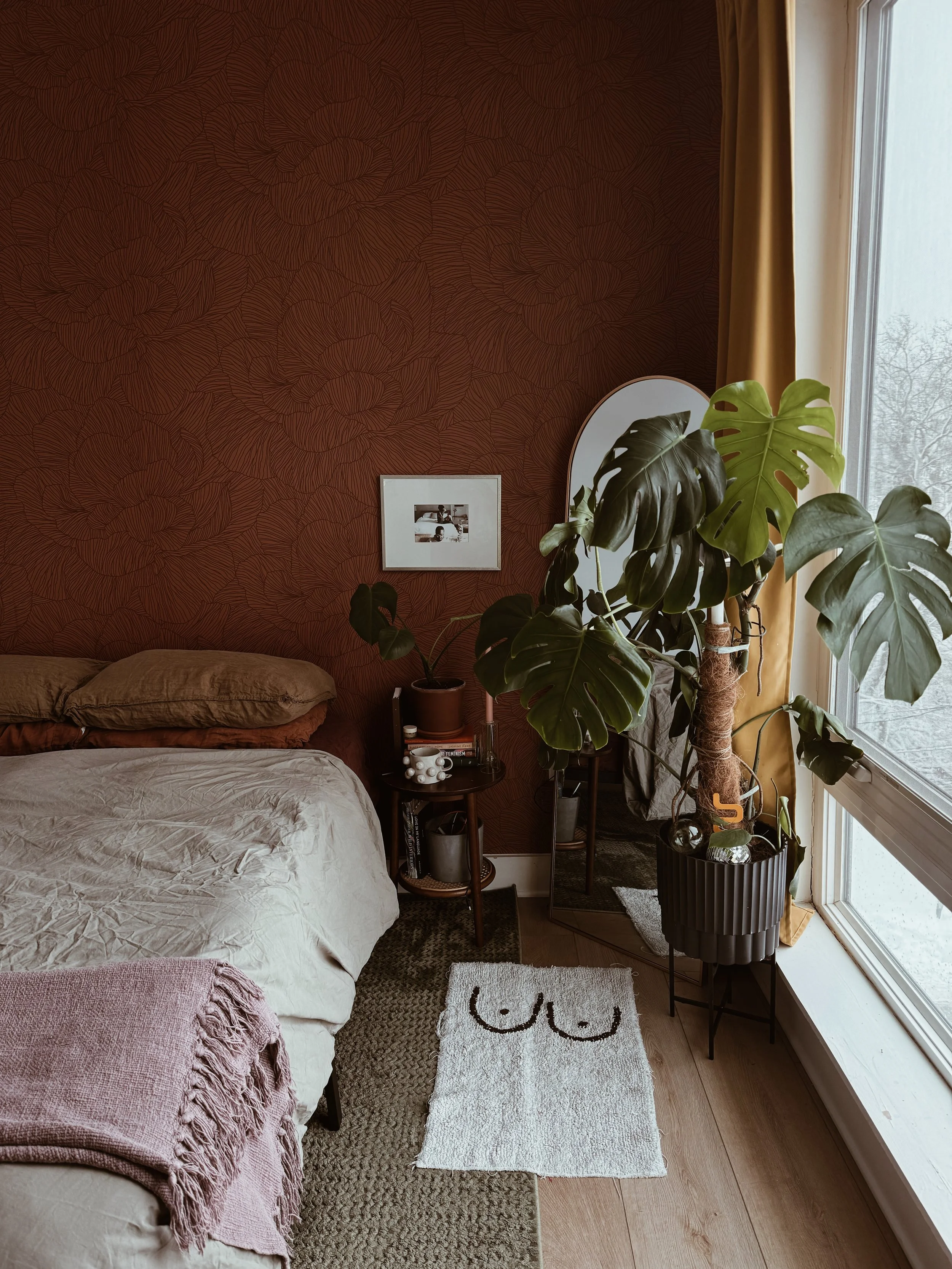 A cozy bedroom corner with a bed, side table, large monstera plant, mirror, and window with yellow curtains.