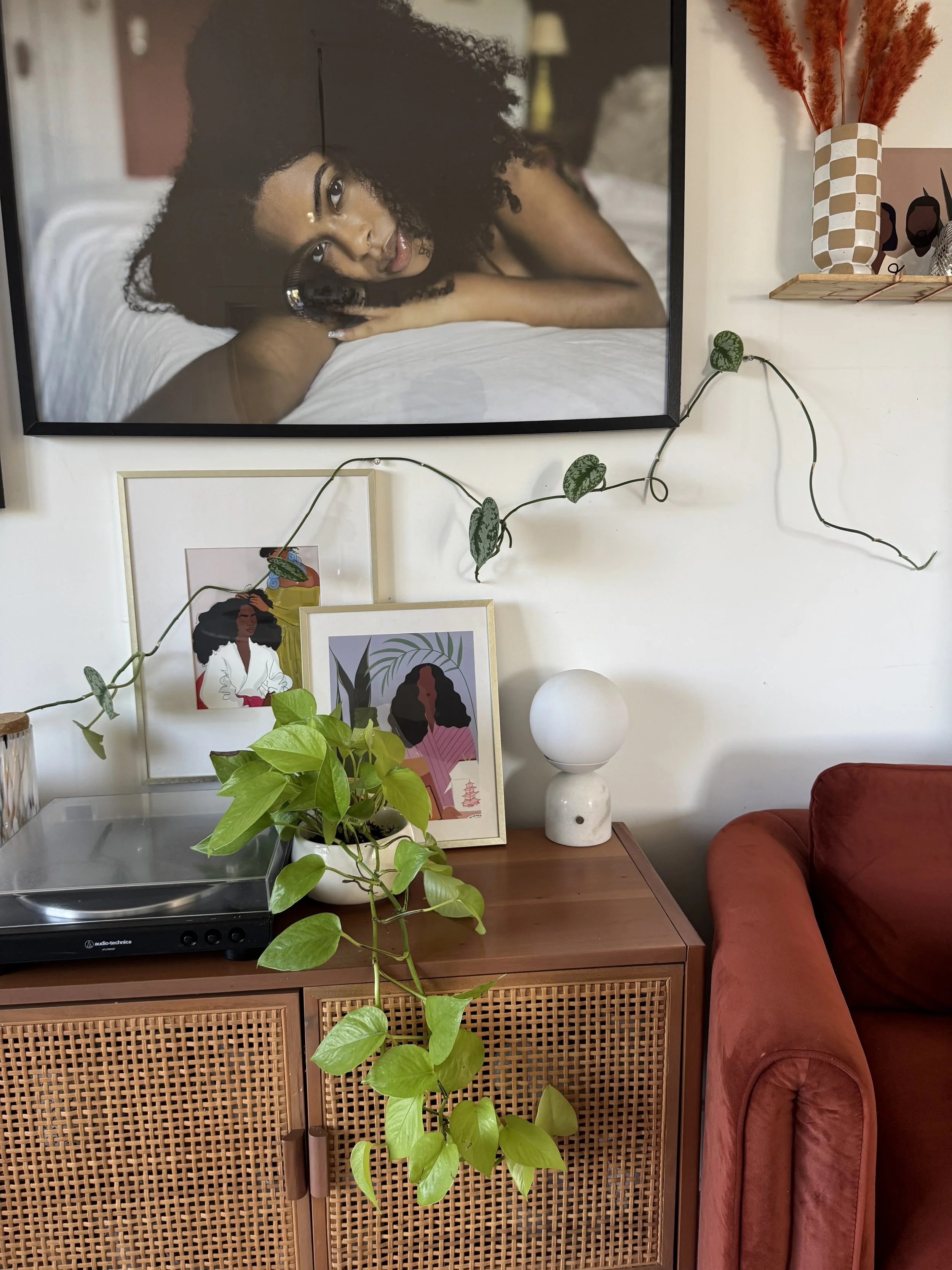 Living room decor with a framed photograph of a woman with curly hair lying on a bed, a potted plant on a wooden cabinet, two framed artwork prints, a white spherical lamp on a marble base, and a reddish-brown sofa.