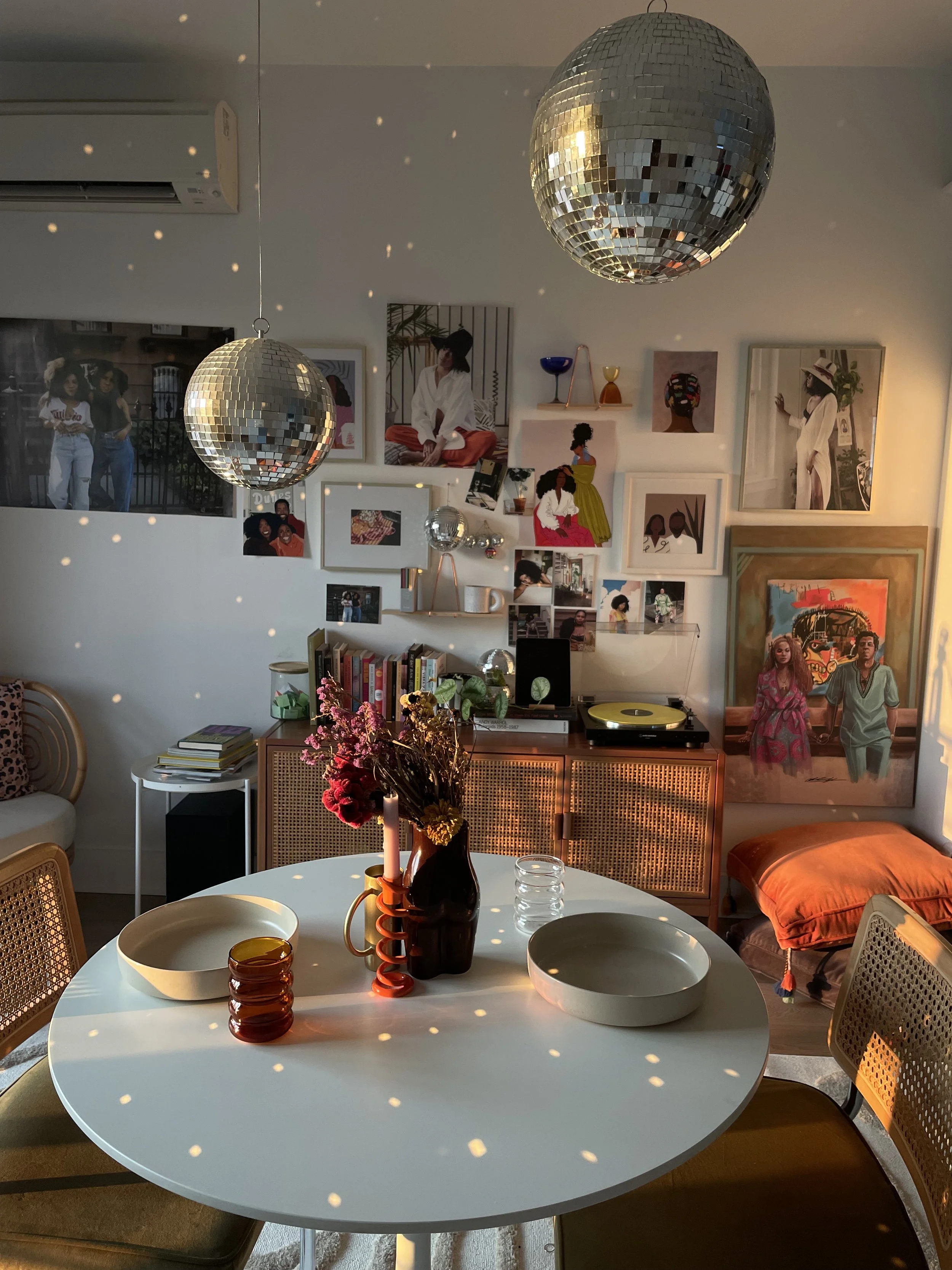 A dining room with sunlight creating disco ball reflections on the ceiling and walls. There is a round white table with a vase of flowers, candles, and bowls. The background features wall art, books, and a record player. Two disco balls hang from the