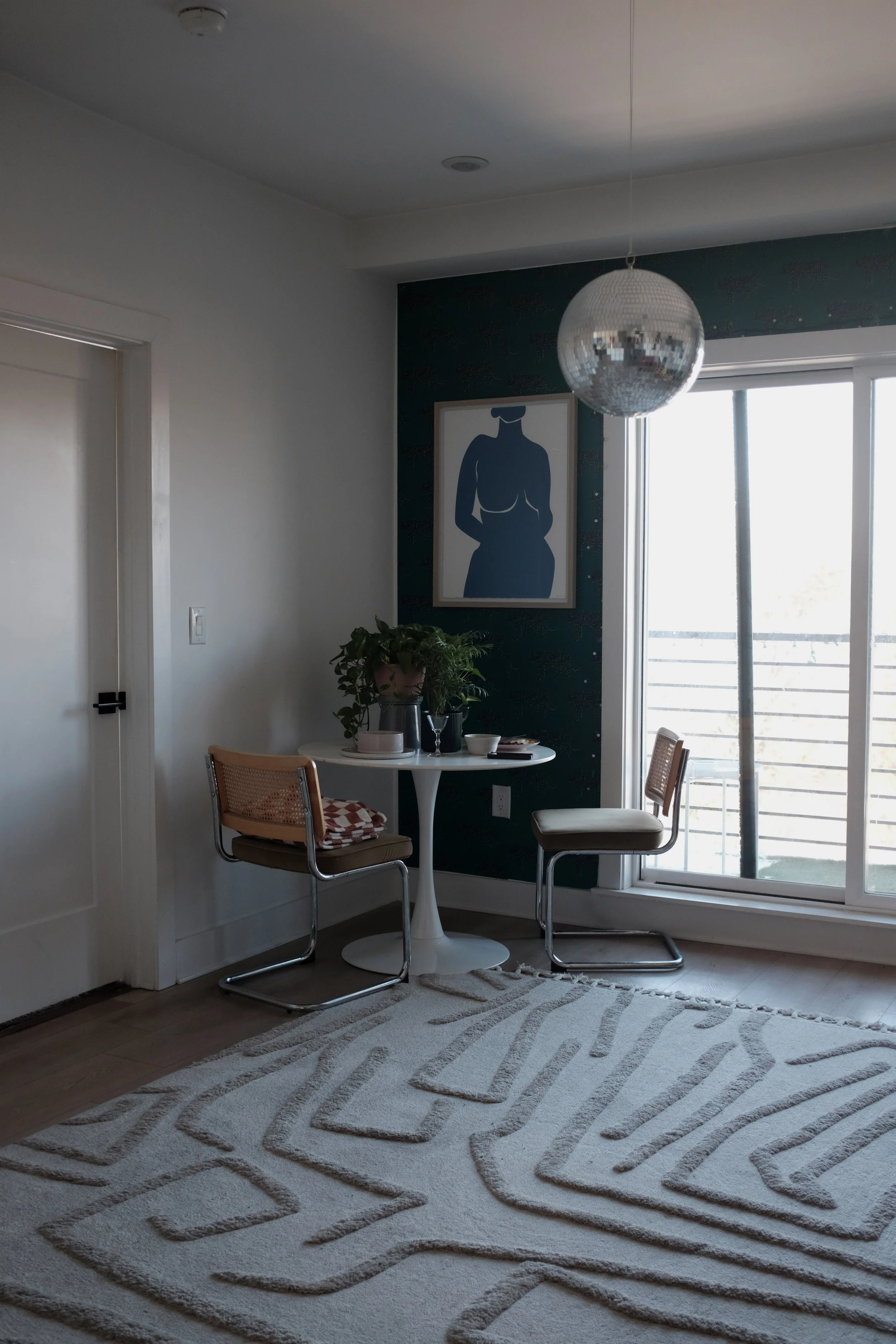 Interior view of a cozy corner with a small round white table, two chairs, a potted plant, a wall art piece, and a sliding glass door leading to a balcony with a mirrored disco ball hanging from the ceiling.