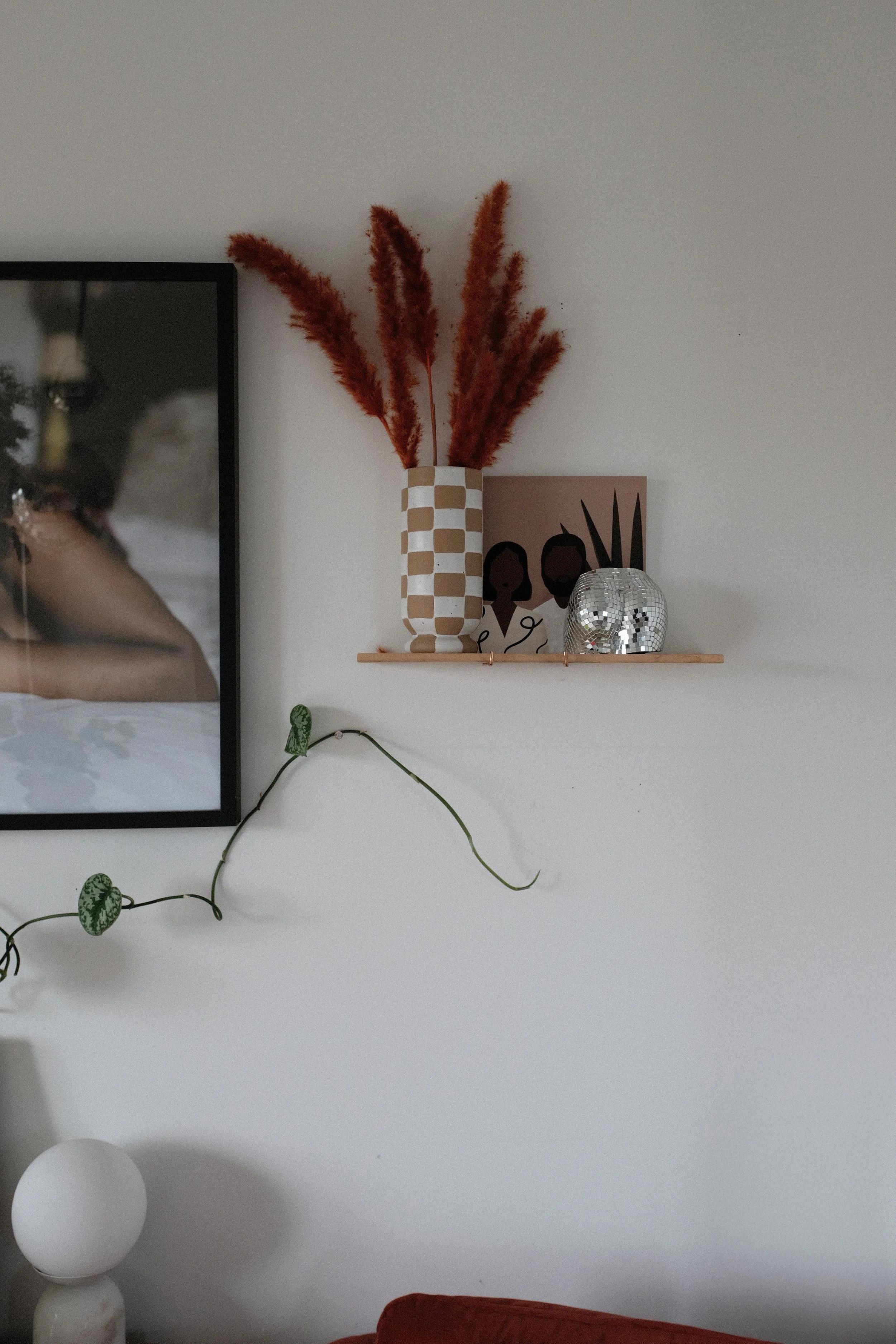 Decorative shelf with a checkered vase holding reddish-brown dried pampas grass, a portrait artwork of two women, a small disco ball, and a metallic pumpkin sculpture. Part of a framed photograph of a person is visible on the left.