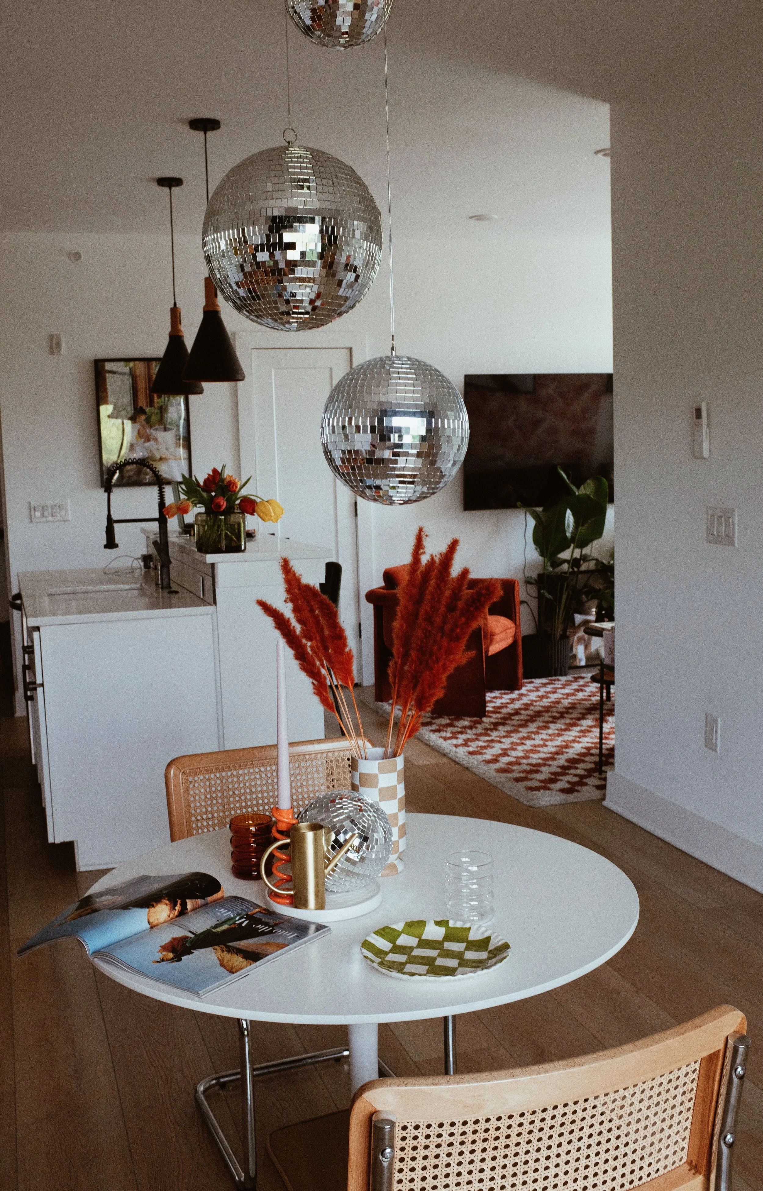 A dining area in a contemporary home featuring a round white table with a decorative vase of orange and pink pampas grass, an open magazine, a green and white checkered plate, and glasses. Overhead, two disco ball pendant lights hang, and in the back