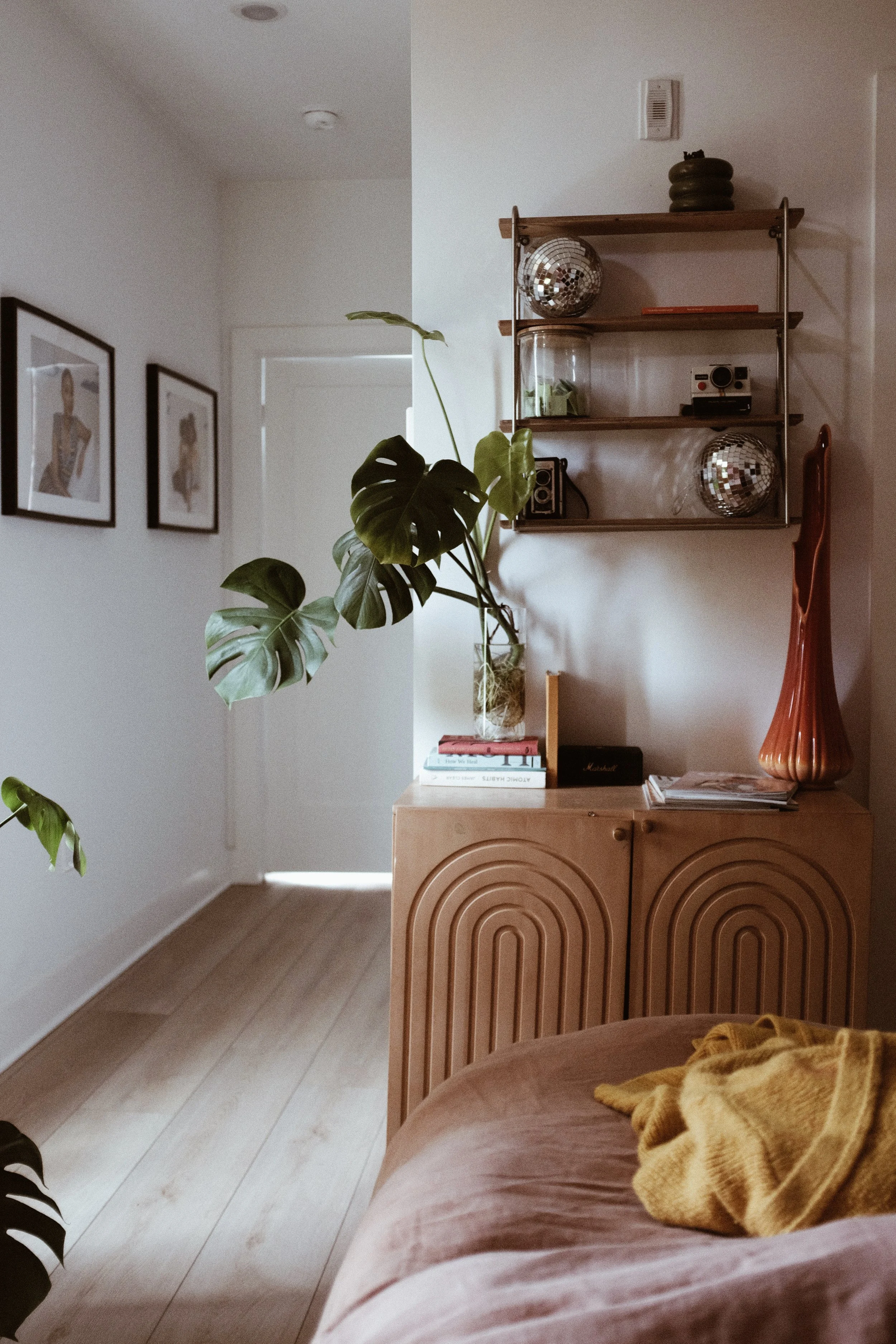 Living room with a beige cabinet decorated with books, a yellowcloth, and a large pink vase, against a white wall with a wall shelf holding disco balls, candles, and cameras, with two potted plants, and three framed pictures on the adjacent wall.