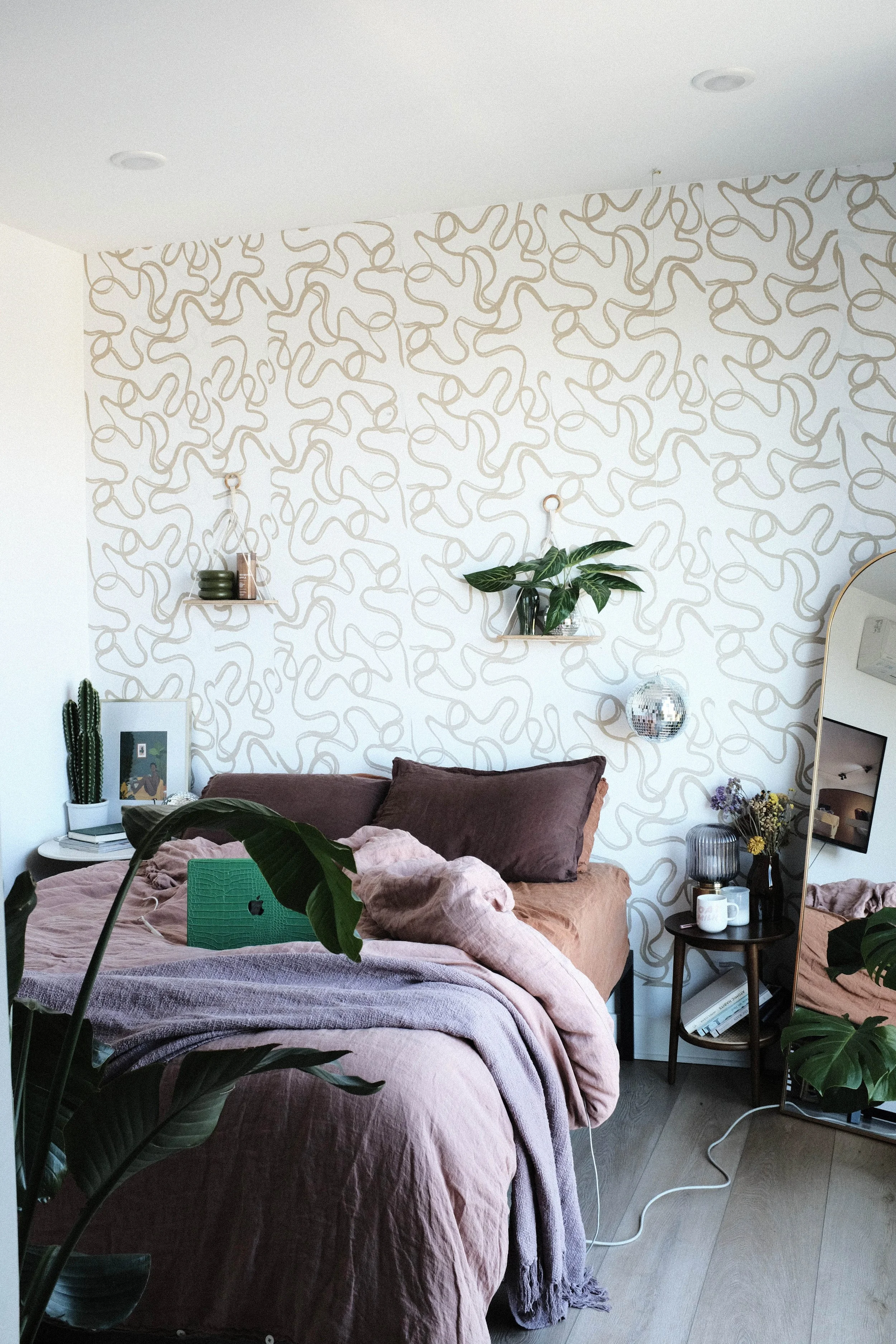 Cozy bedroom with pink and brown bedding, green plants, wall with abstract beige line art pattern, small side table with books, mug, and flowers, standing mirror, and various decorative items.