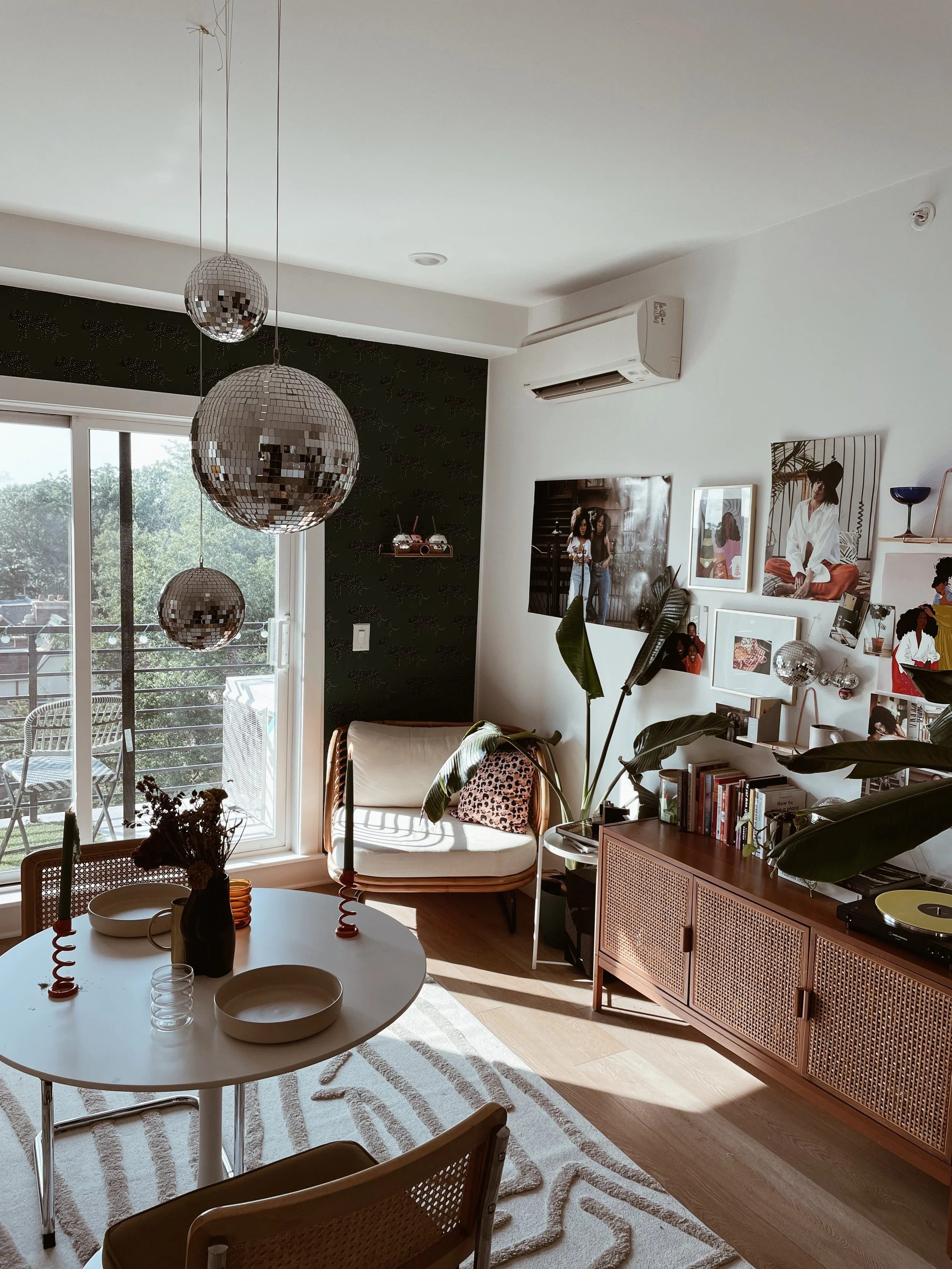 A living room with hanging disco ball lights, a round dining table with bowls and candles, a white upholstered chair with a leopard print cushion, a wooden sideboard with books and plants, and a gallery wall of framed photos. Large windows and a slid