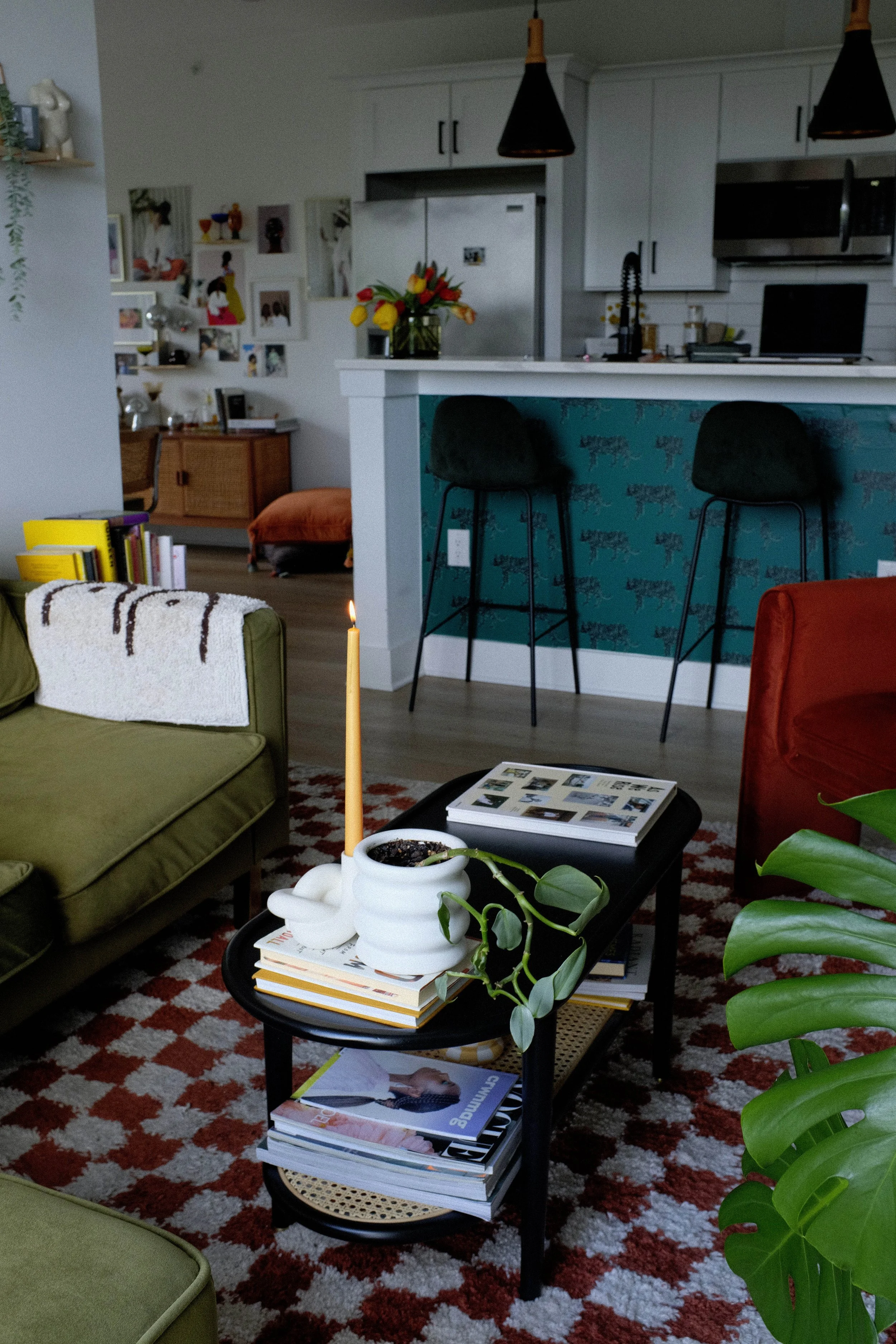 Living room with a black coffee table topped with magazines, a white ceramic planter with a trailing plant, and a single lit taper candle. Green and red armchairs, colorful patterned rug, and a kitchen area in the background with black bar stools, wh