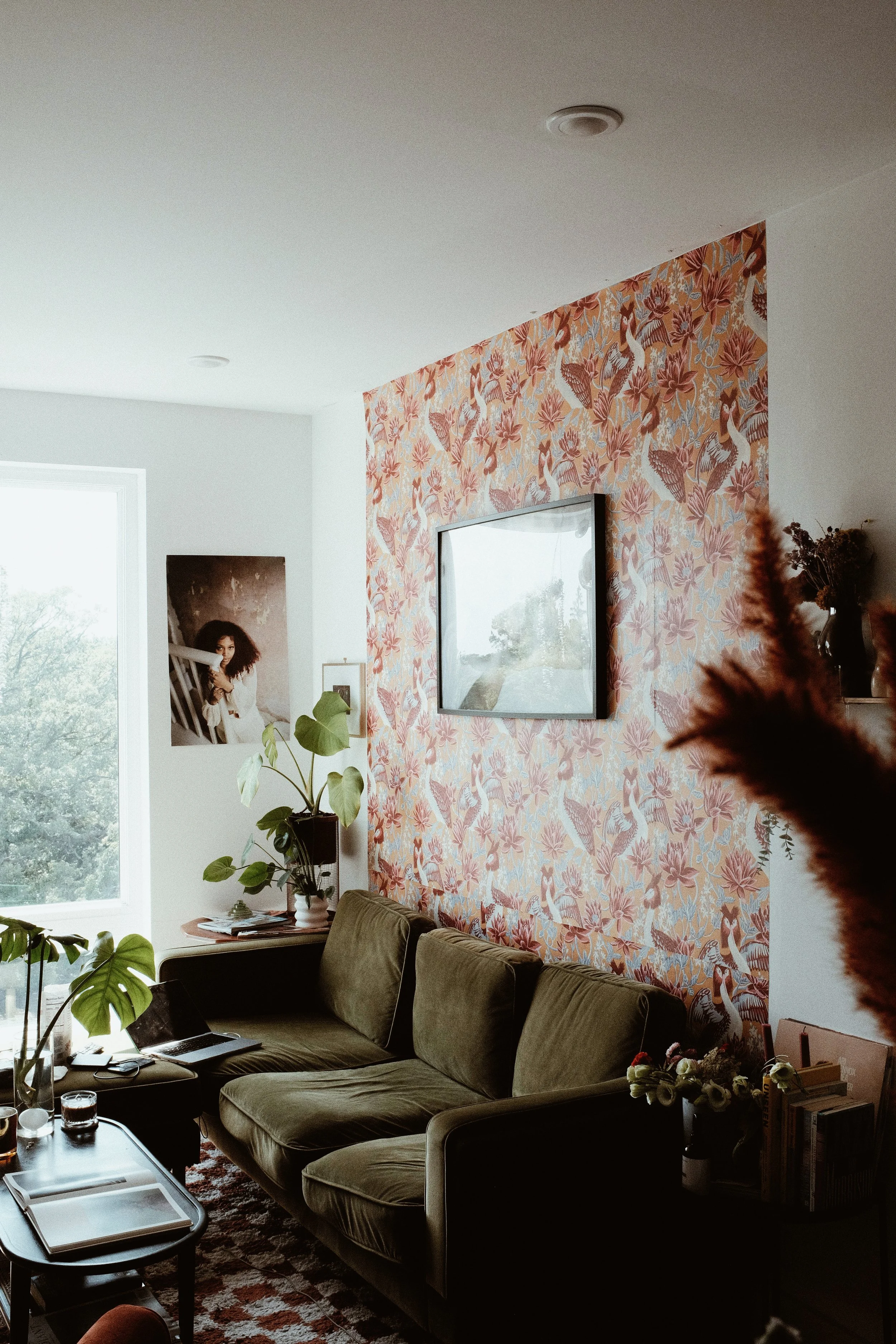 Living room with a green velvet sofa, patterned wallpaper, wall-mounted TV, large window, potted plants, framed photo, and decorative objects.