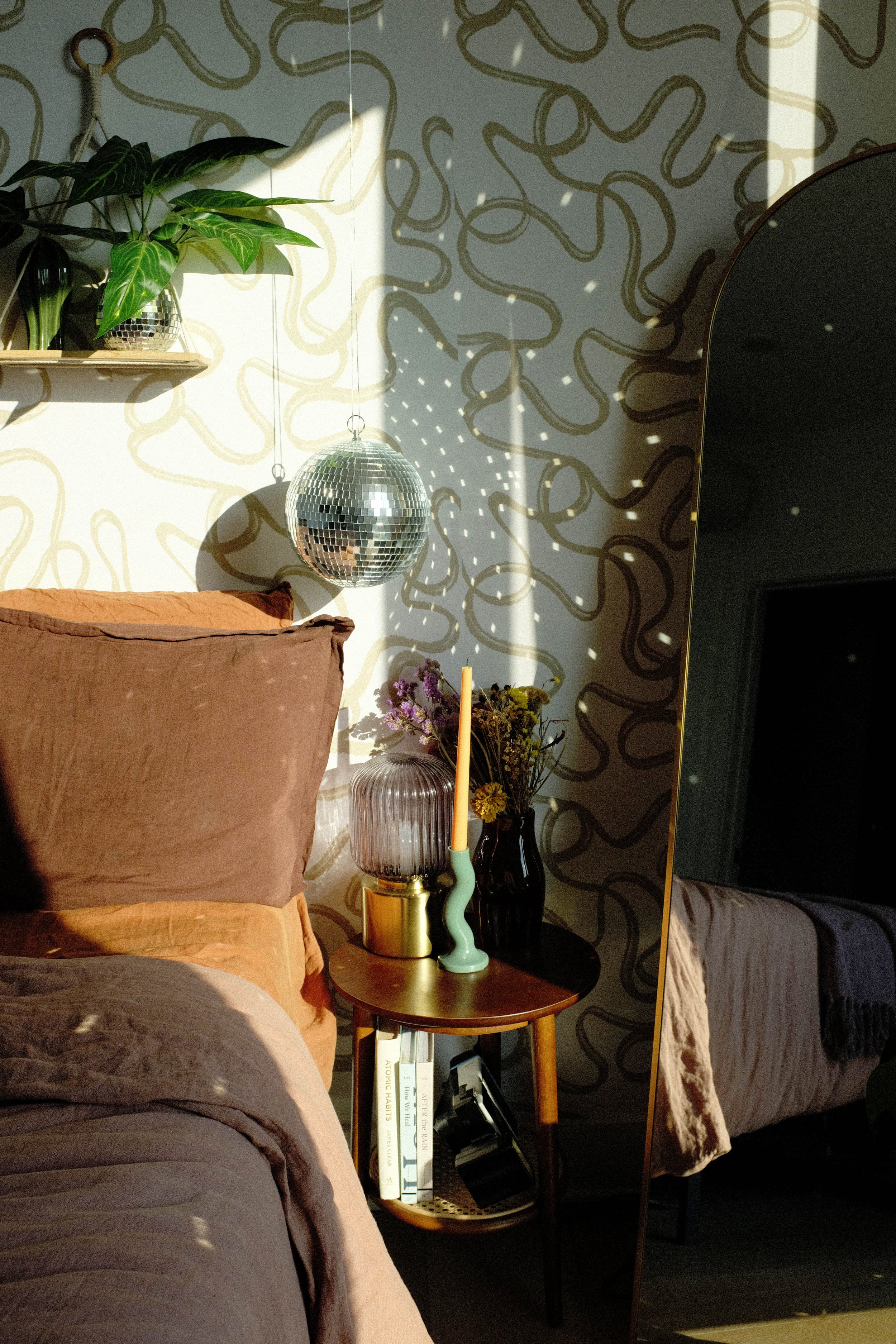 A cozy bedroom corner with a bed, a side table with vases and a candle, a mirror, and a wall decorated with abstract gold patterns, illuminated by sunlight and a disco ball.