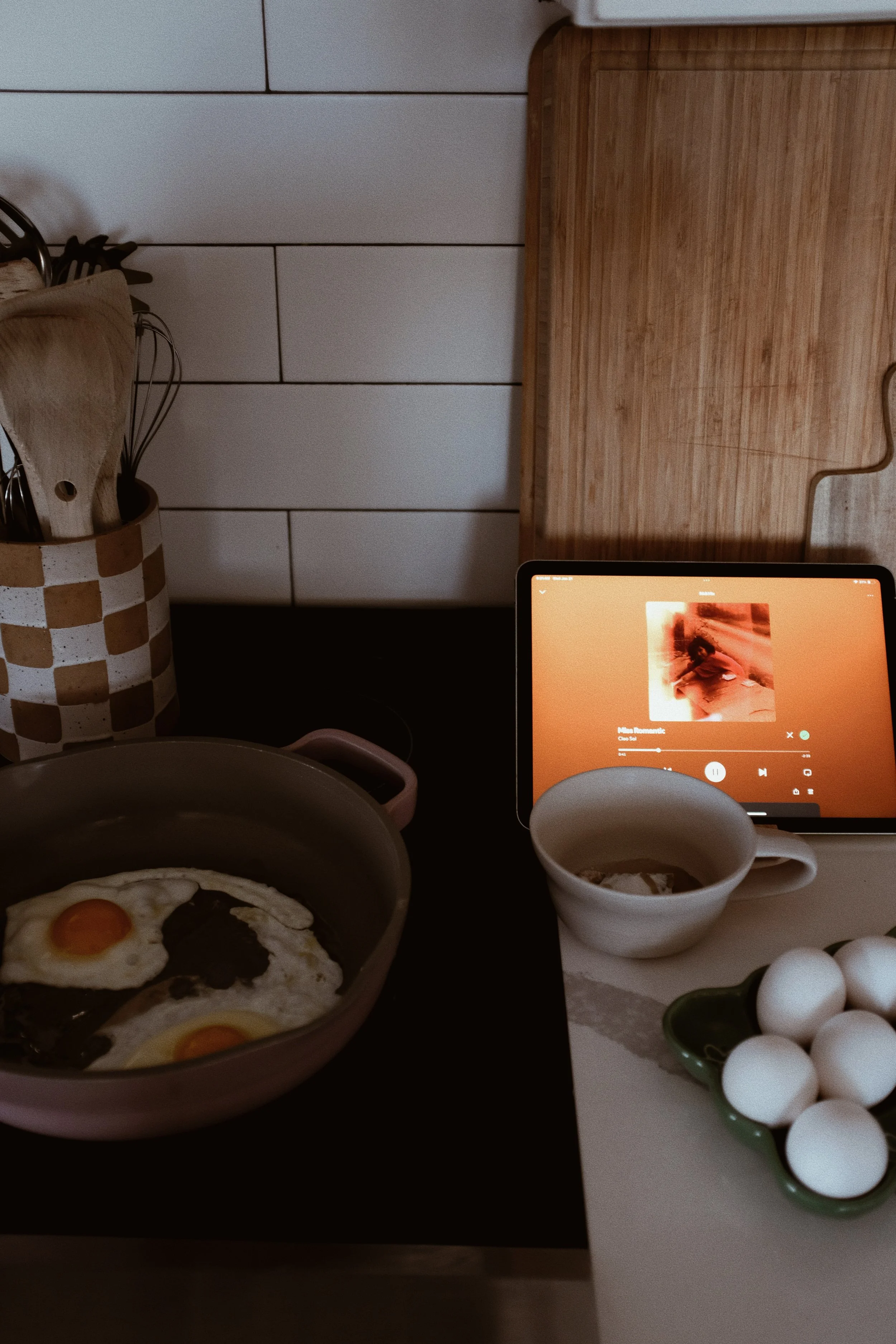A kitchen countertop with a pot of fried eggs, an iPad playing music, an empty white mug, a bowl of eggs, and various kitchen utensils in a container. The background features a wooden cutting board and white tiled wall.