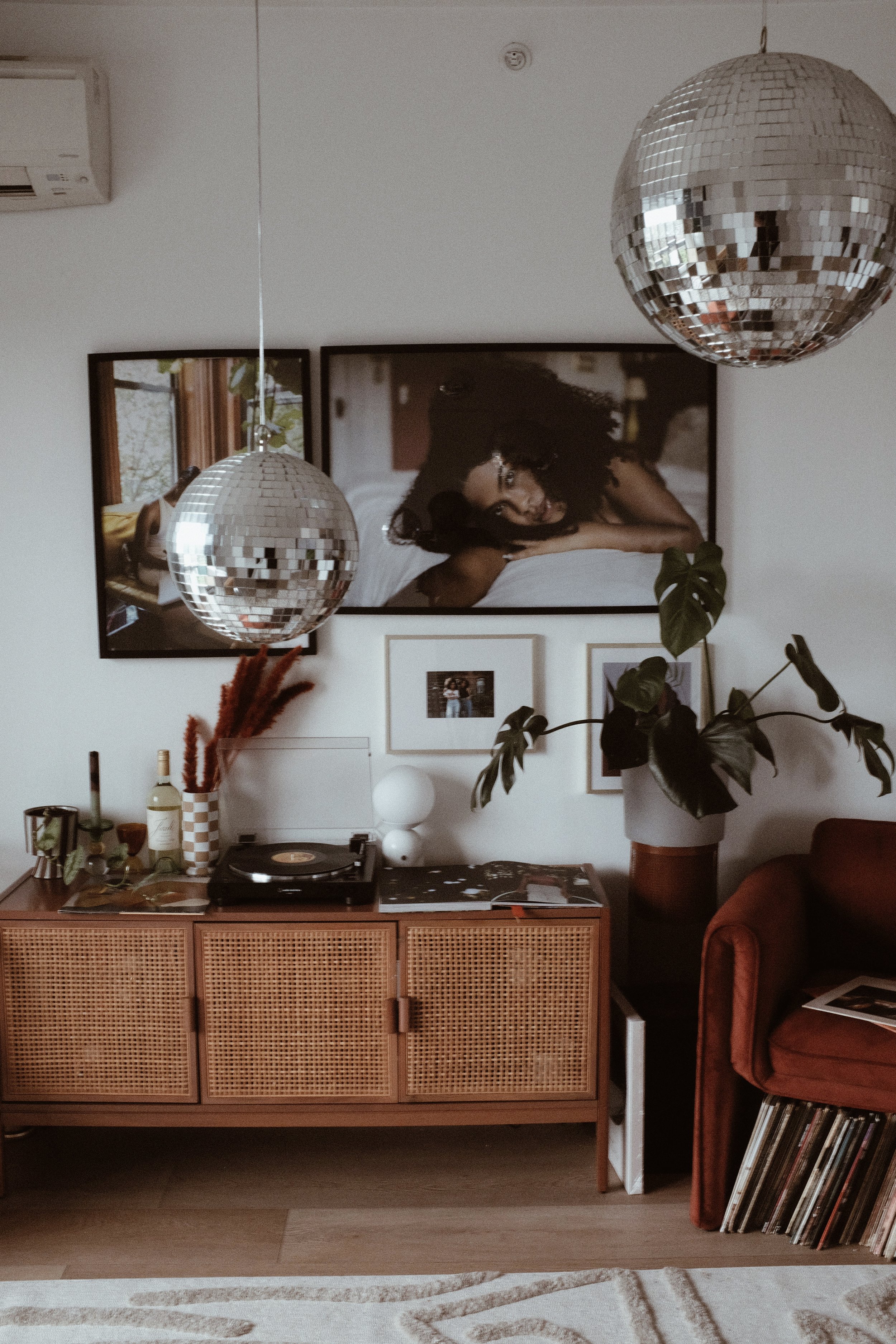 Living room with framed photographs on the wall, two disco balls hanging from the ceiling, a vintage wooden sideboard with a record player, bottles, and decor, a large potted plant, a rust-colored armchair with magazines or photographs on it, and a p
