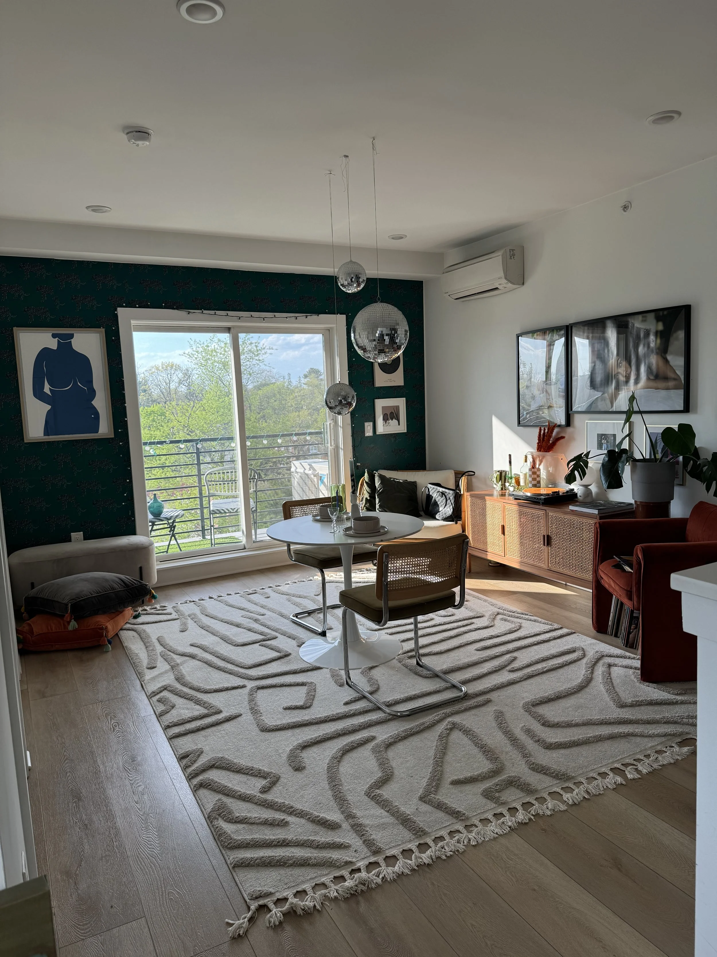 Modern living room with a sliding glass door leading to a balcony, featuring a round table with two chairs, a white patterned rug, a sideboard with plants and decor, framed artwork on the walls, and pendant lights hanging from the ceiling.