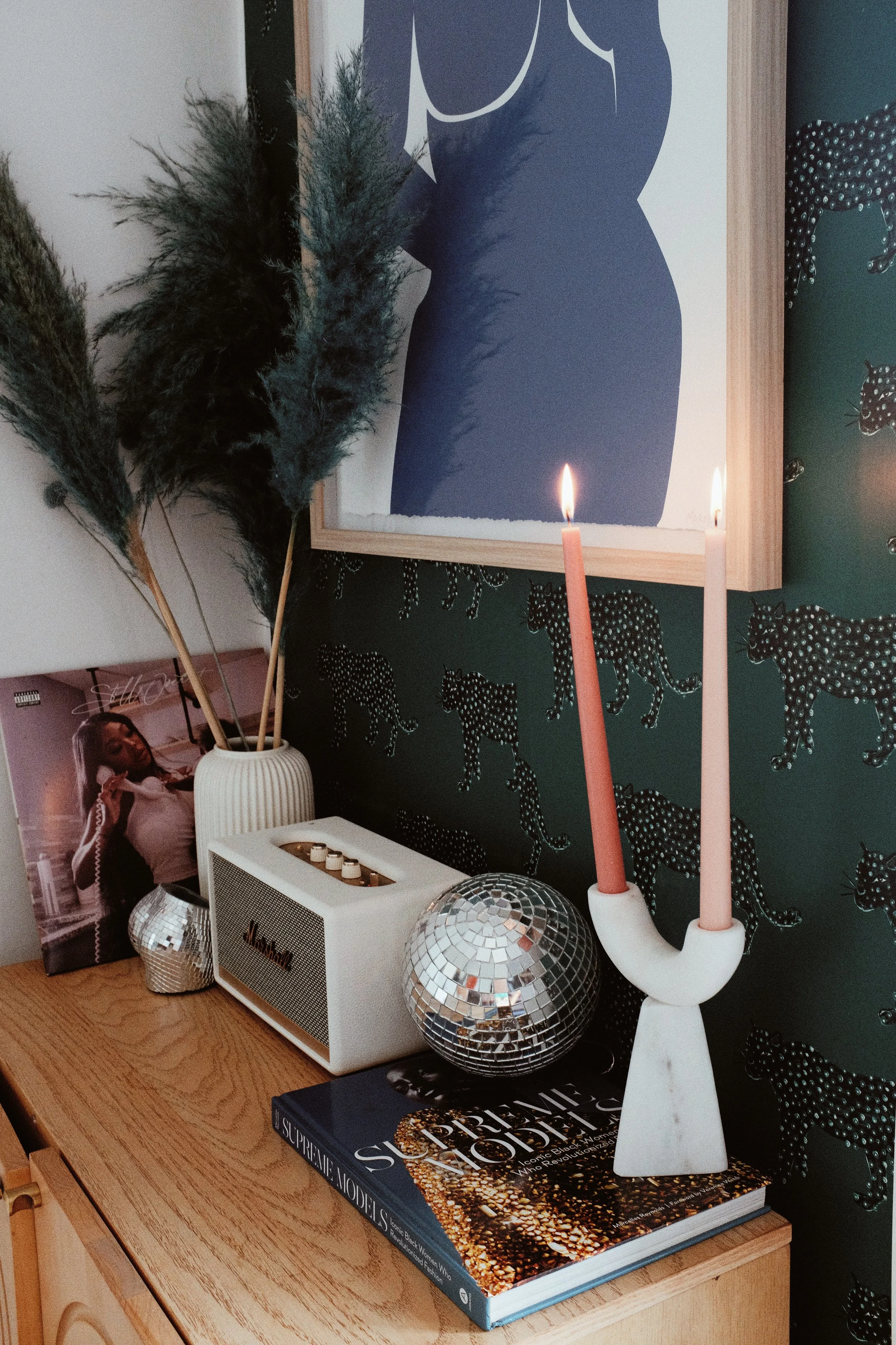 Decorative tabletop arrangement on a wooden surface, featuring a small disco ball, a white radio, a framed art piece, a magazine titled 'Supreme Models,' a white vase with tall pampas grass, and a white candle holder with two tall candles—one pink an