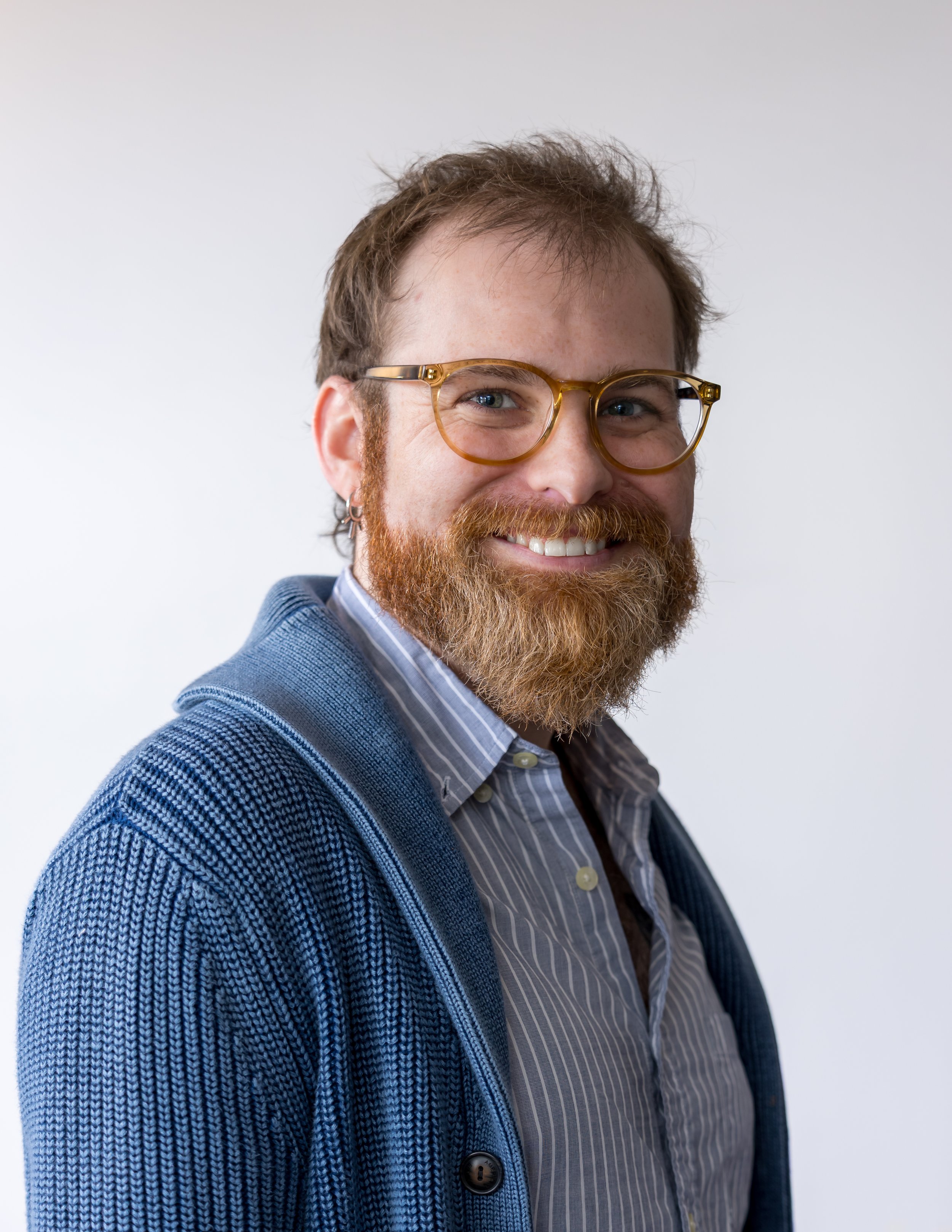 Close-up of a smiling man with brown hair, glasses, and a beard, wearing a blue cardigan over a striped shirt, against a plain white background.