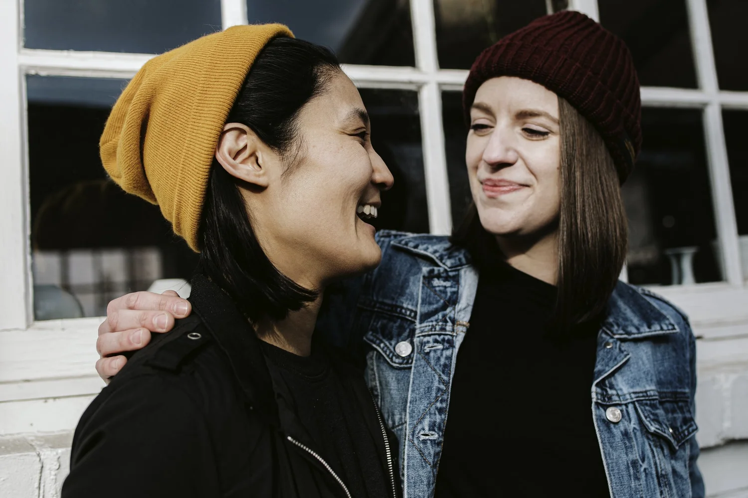 Two people in knit hats smiling at one another, with an arm around a shoulder.