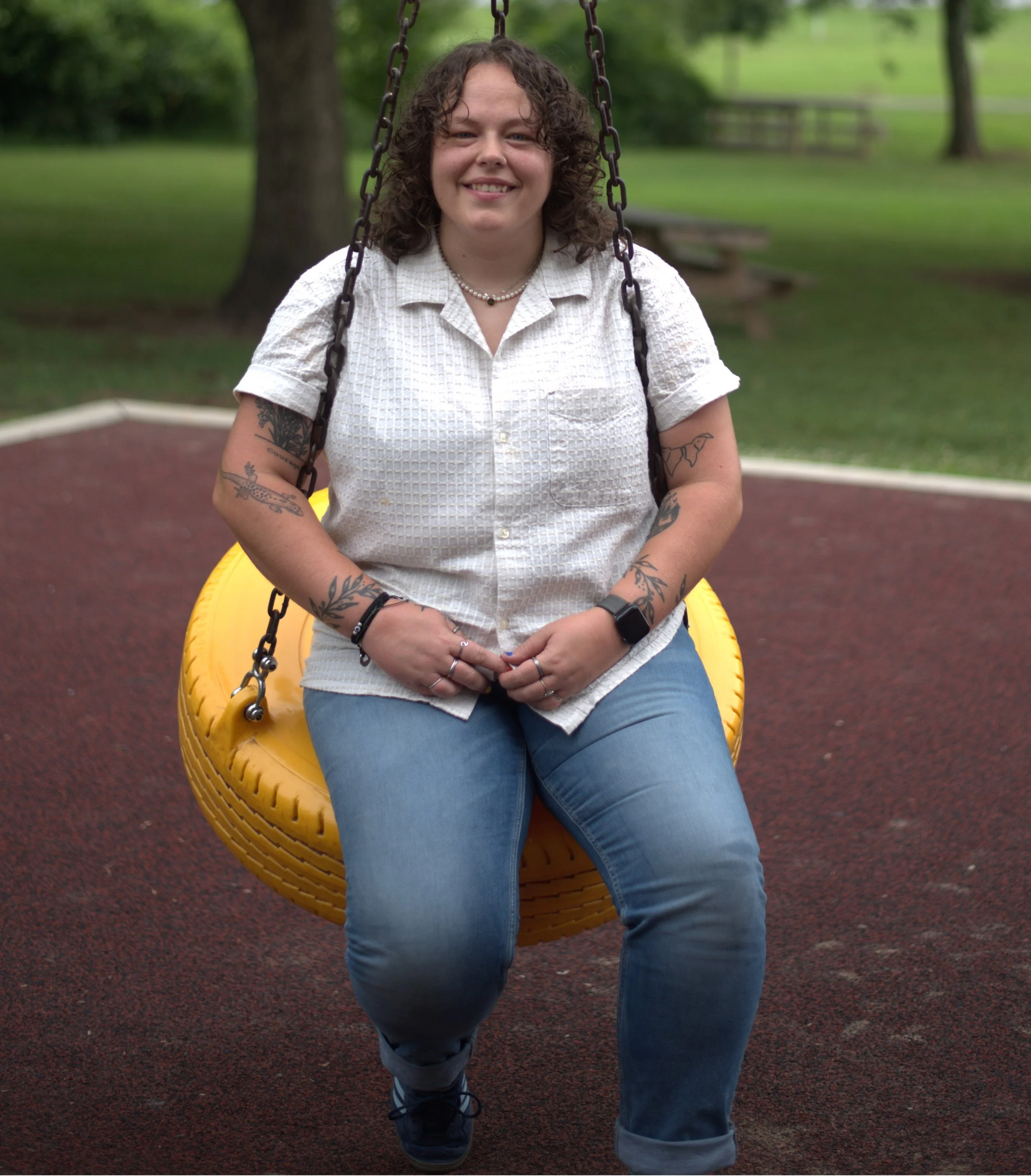A woman with tattoos sitting on a yellow swing at a park, smiling, with green trees in the background.
