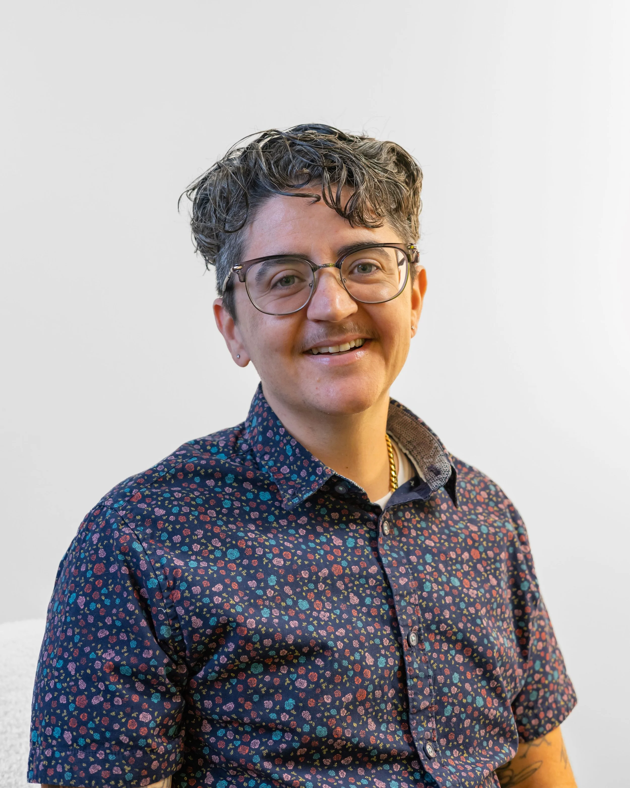 Close-up of a smiling woman wearing glasses and a dark floral shirt, with a plain white background.