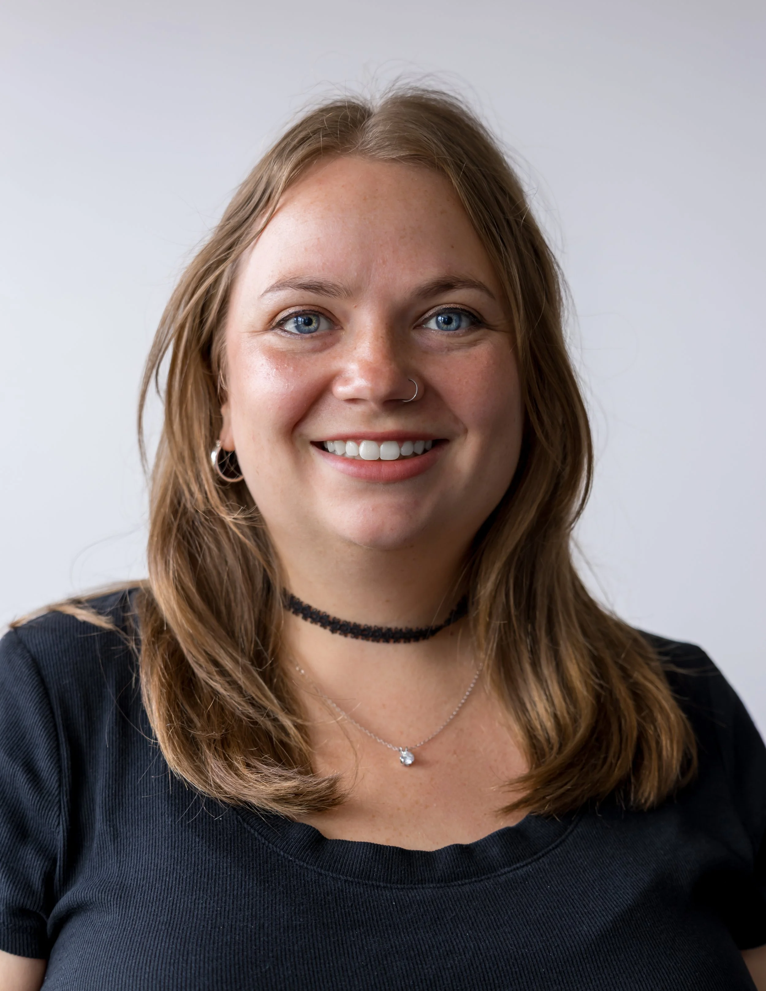 A woman with shoulder-length light brown hair, blue eyes, and a nose piercing, smiling and taking a selfie indoors. She is wearing a black top with a floral patterned cover-up or jacket over it, and a necklace with a small charm.