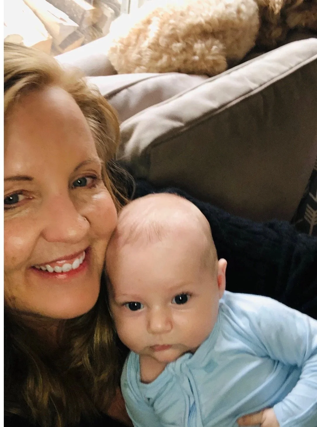 Doula Christina with long, wavy red hair smiling and holding a baby with a bald head and blue eyes. They are close together, indoors, with a sofa and a blanket or pillow in the background.