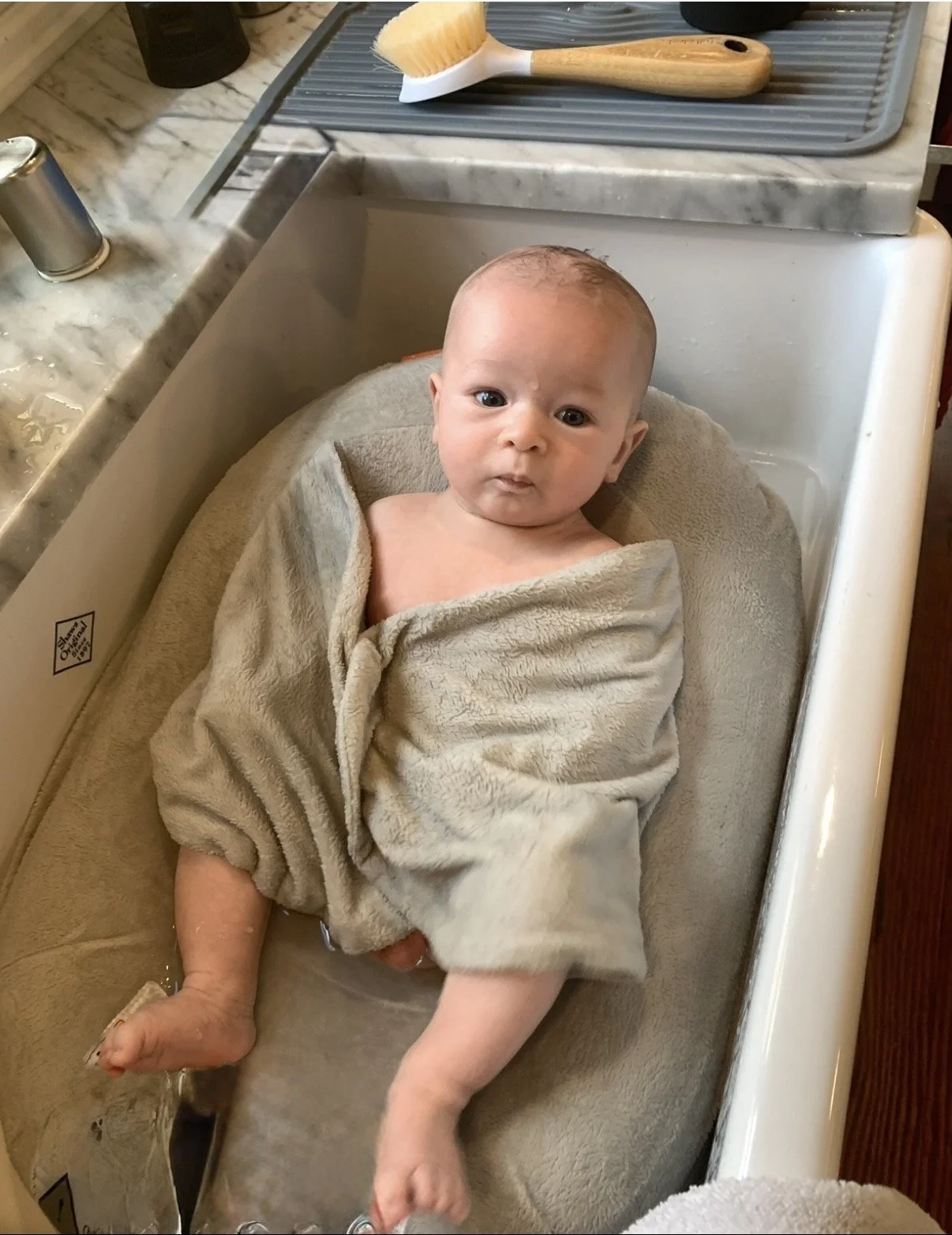 Baby in a beige towel lying in a white kitchen sink with a neutral expression.
