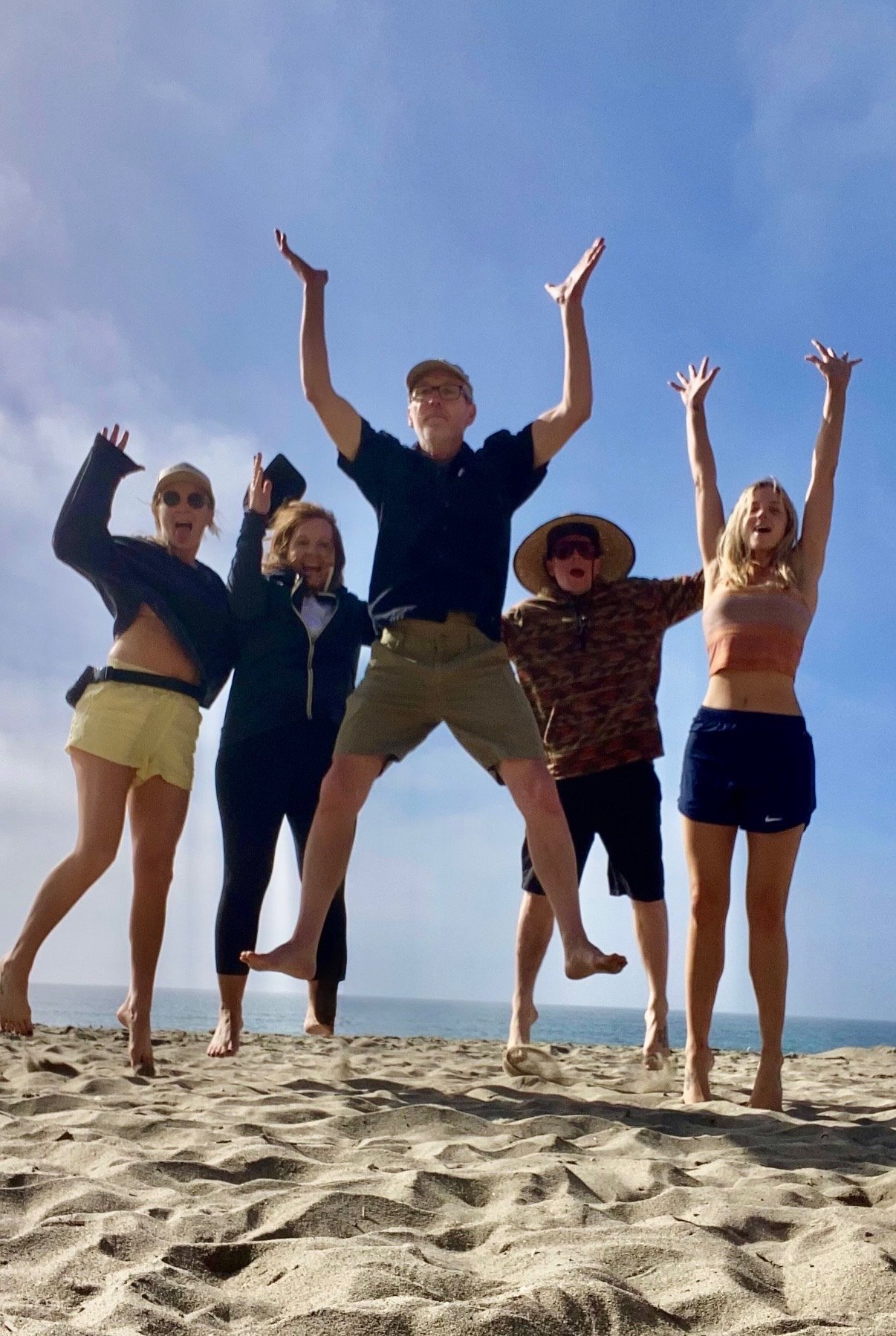 Doula Christina with her family jumping on a sandy beach with the ocean and blue sky in the background.