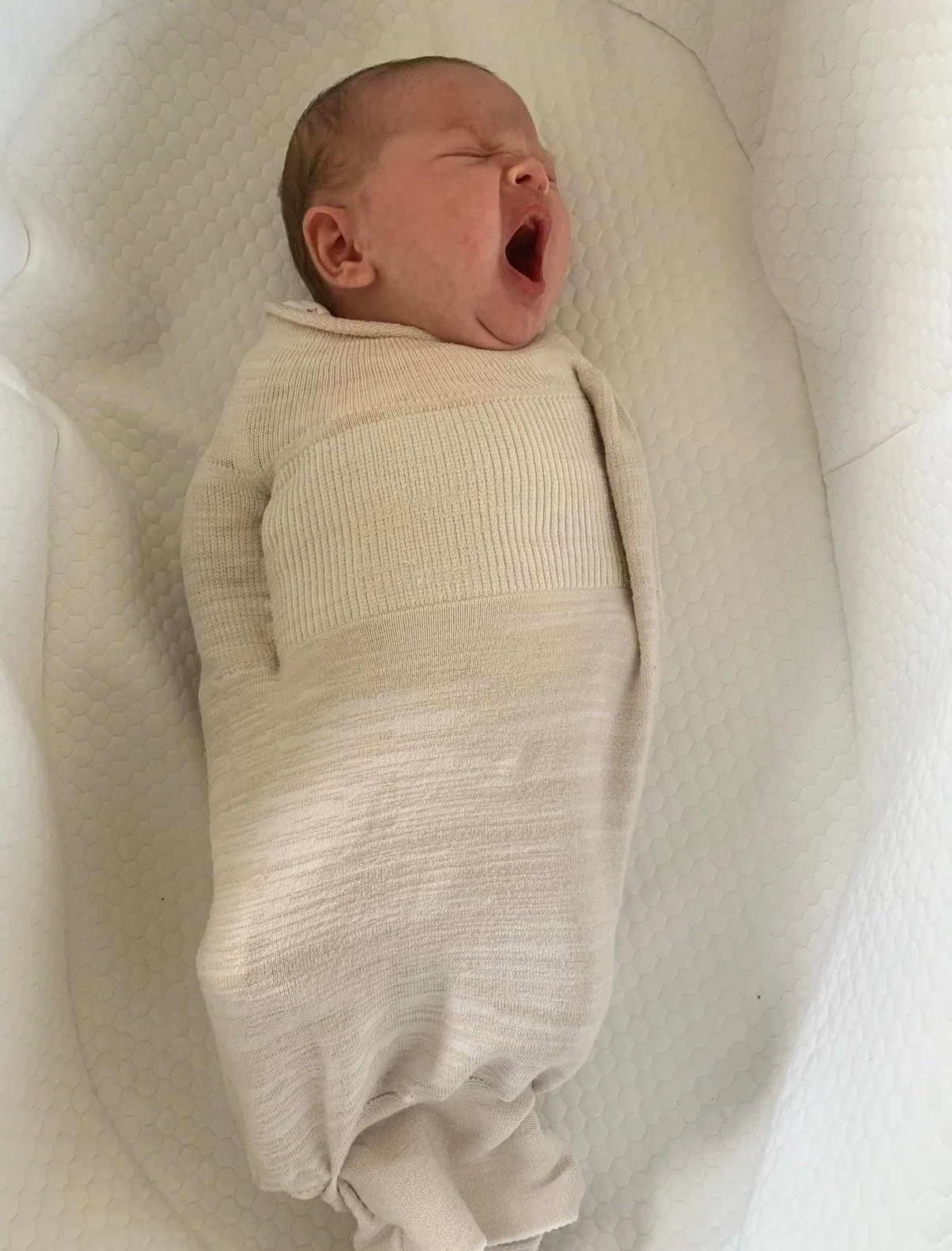 A newborn baby yawning, wrapped in a beige swaddle, lying on a white textured surface.