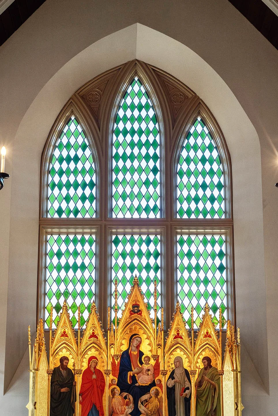 A religious altar with colorful statues of saints and the Virgin Mary holding baby Jesus, set against a large stained glass window with a diamond pattern in shades of green and white.