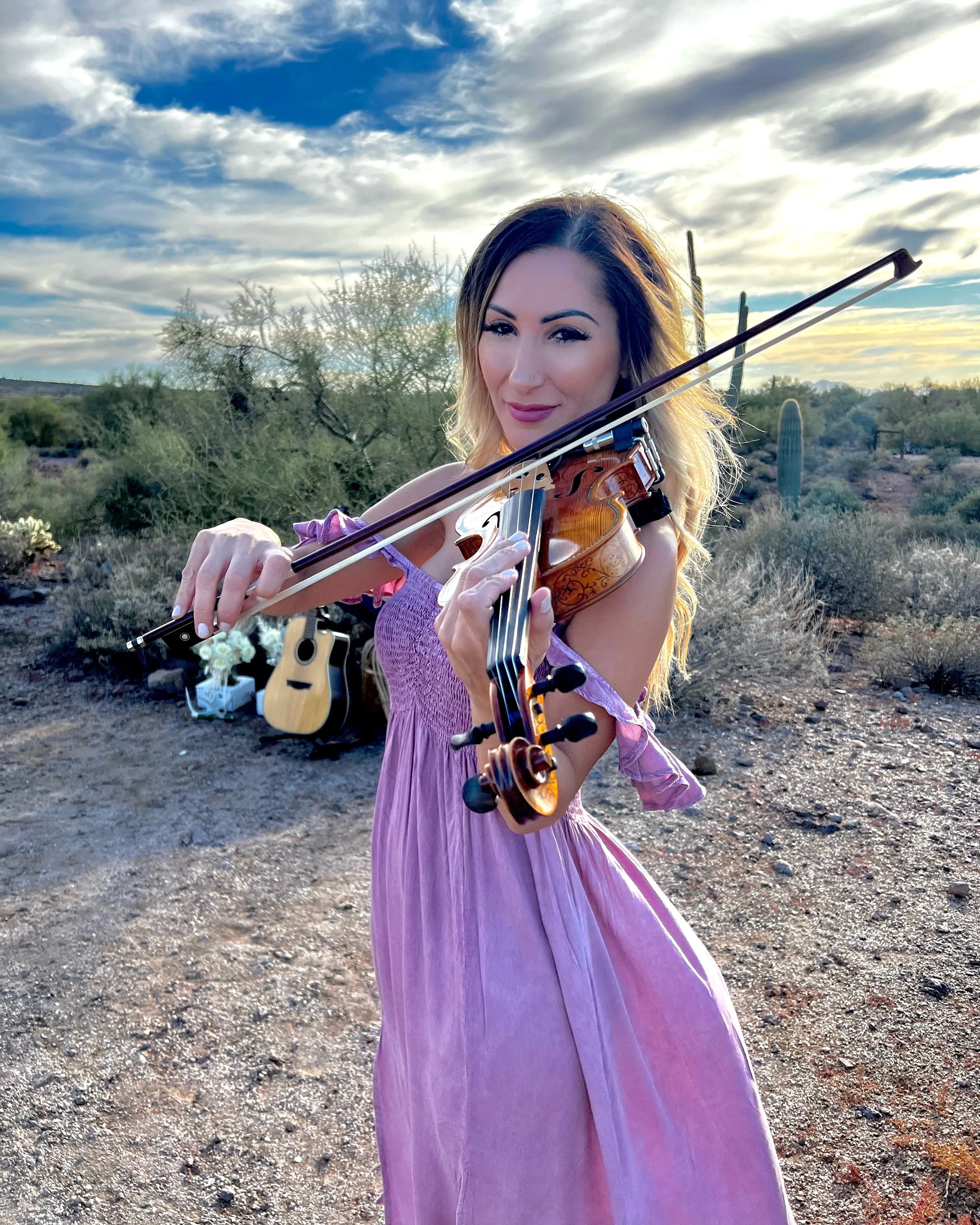 Woman in a pink dress playing a violin outdoors in a desert landscape with cactus and cloudy sky.