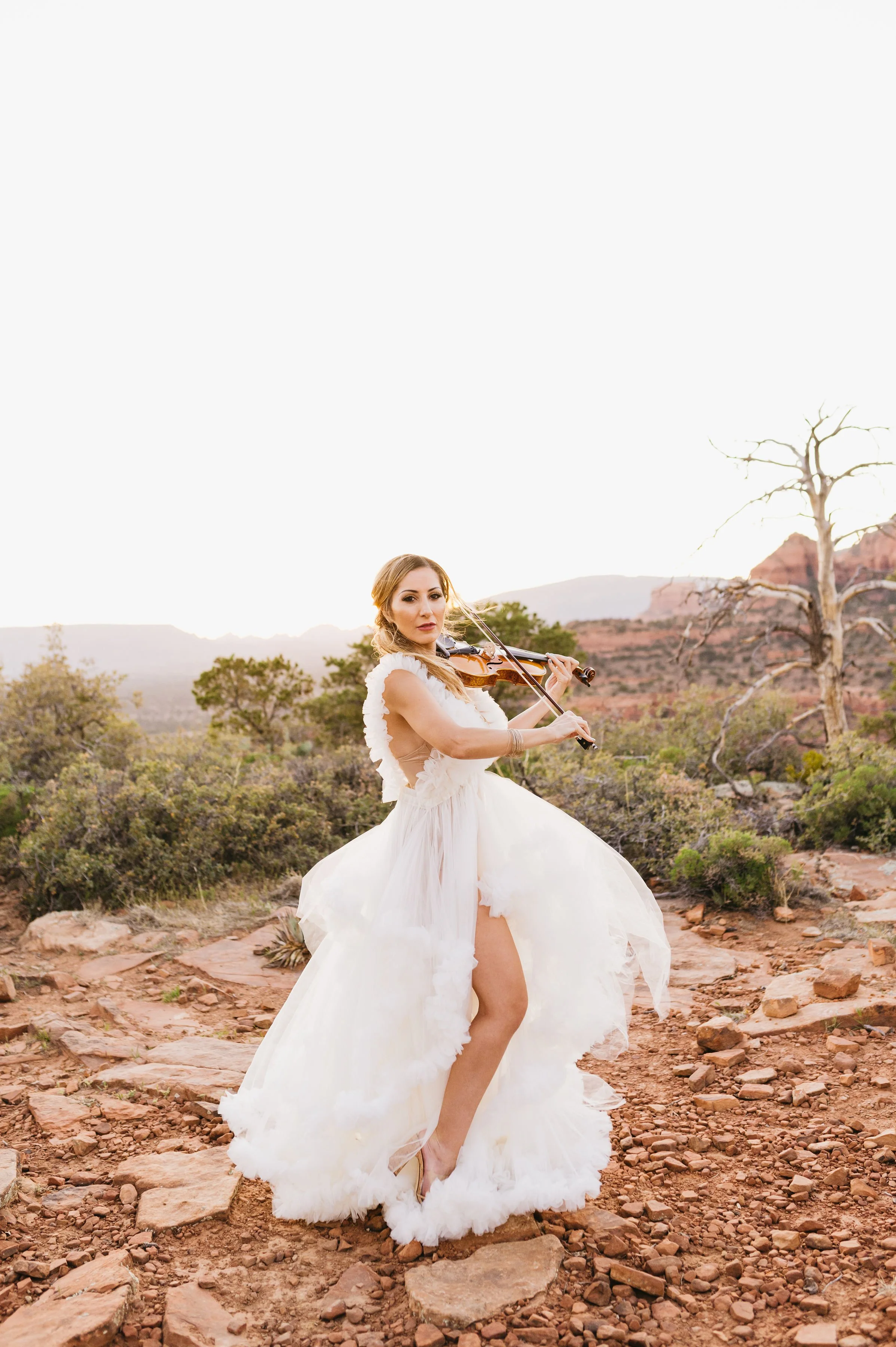 A woman in a flowing white dress plays the violin in a desert landscape with bushes and rocky terrain.