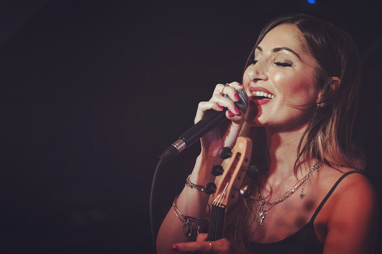 A woman singing into a microphone while holding a guitar, performing onstage with a dark background.