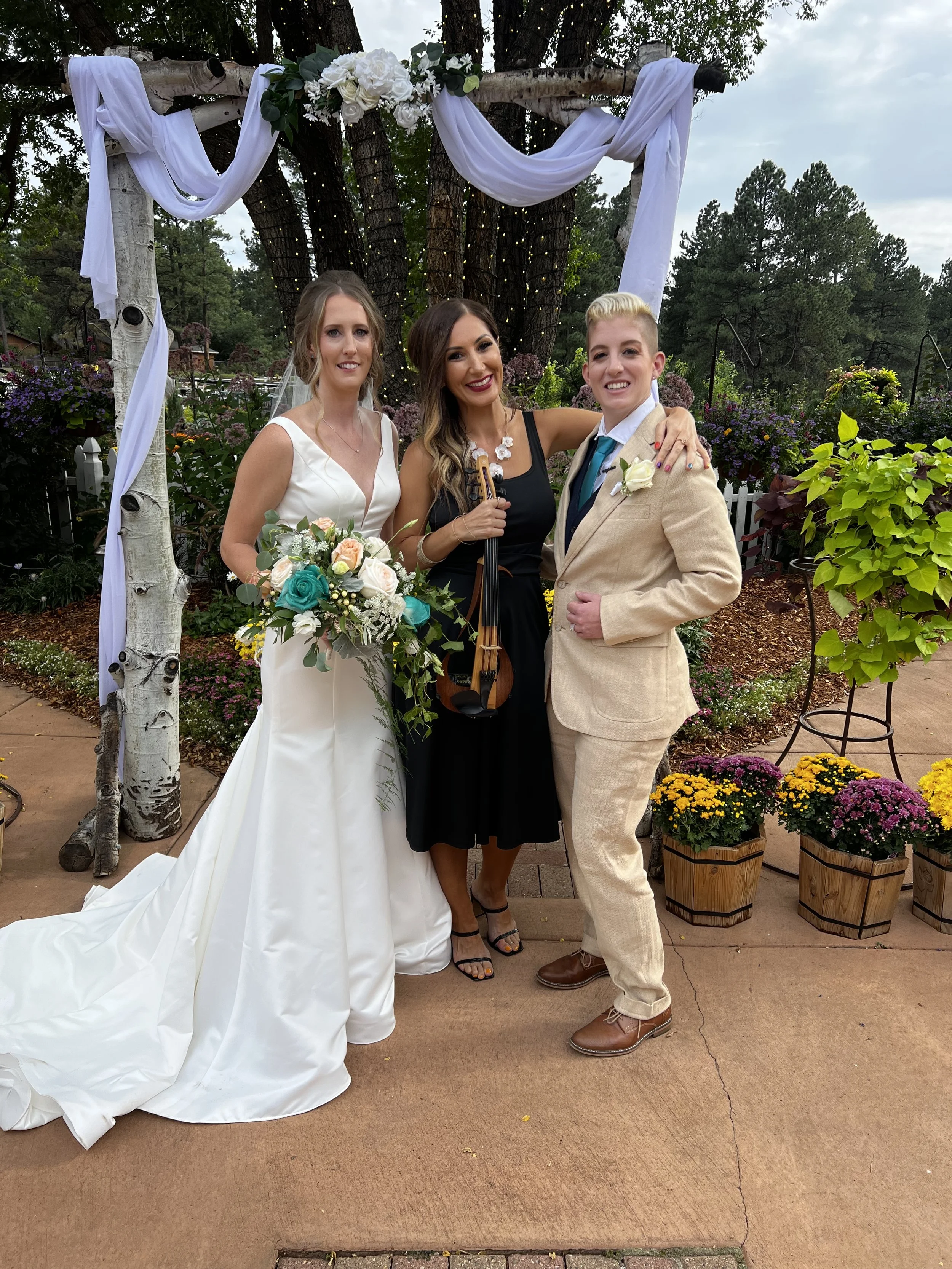A wedding scene with a bride in a white dress holding a bouquet, standing next to a partner in a beige suit. They are with a musician holding a violin, under a decorated wooden arch with white fabric and flowers, surrounded by greenery and flowers.