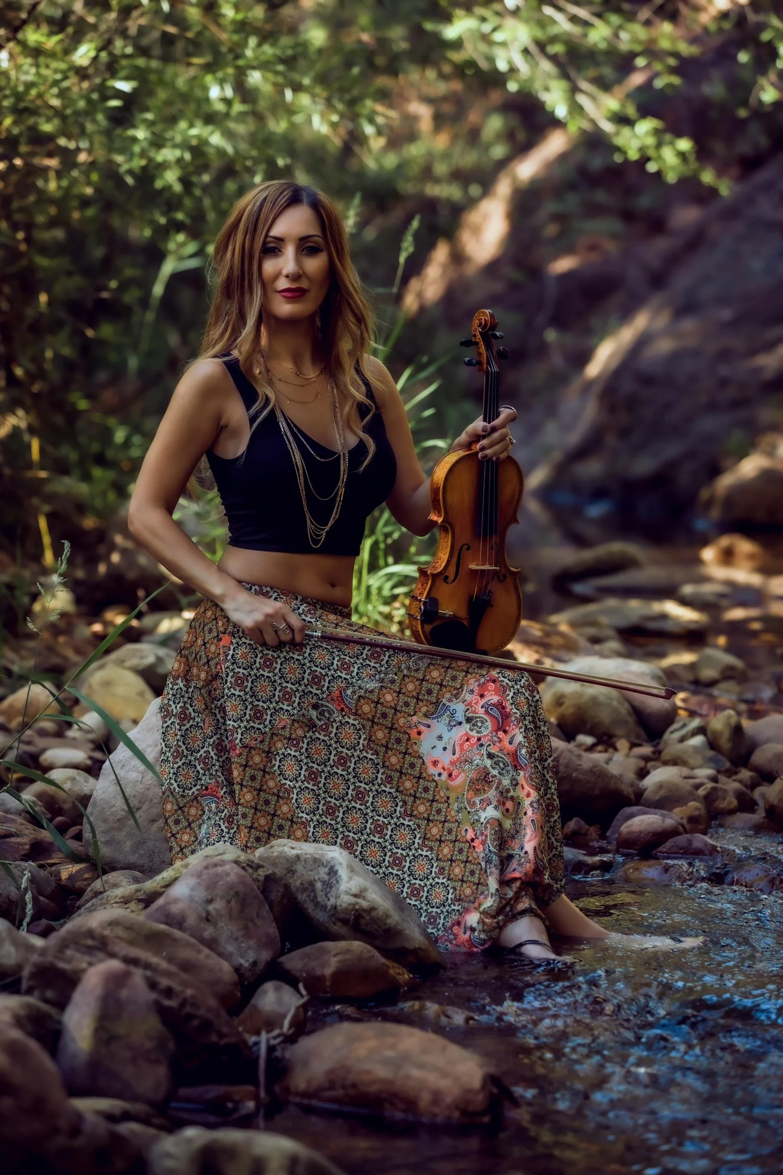 Woman in patterned skirt holding a violin and bow, sitting on rocks in a forest stream, surrounded by greenery.