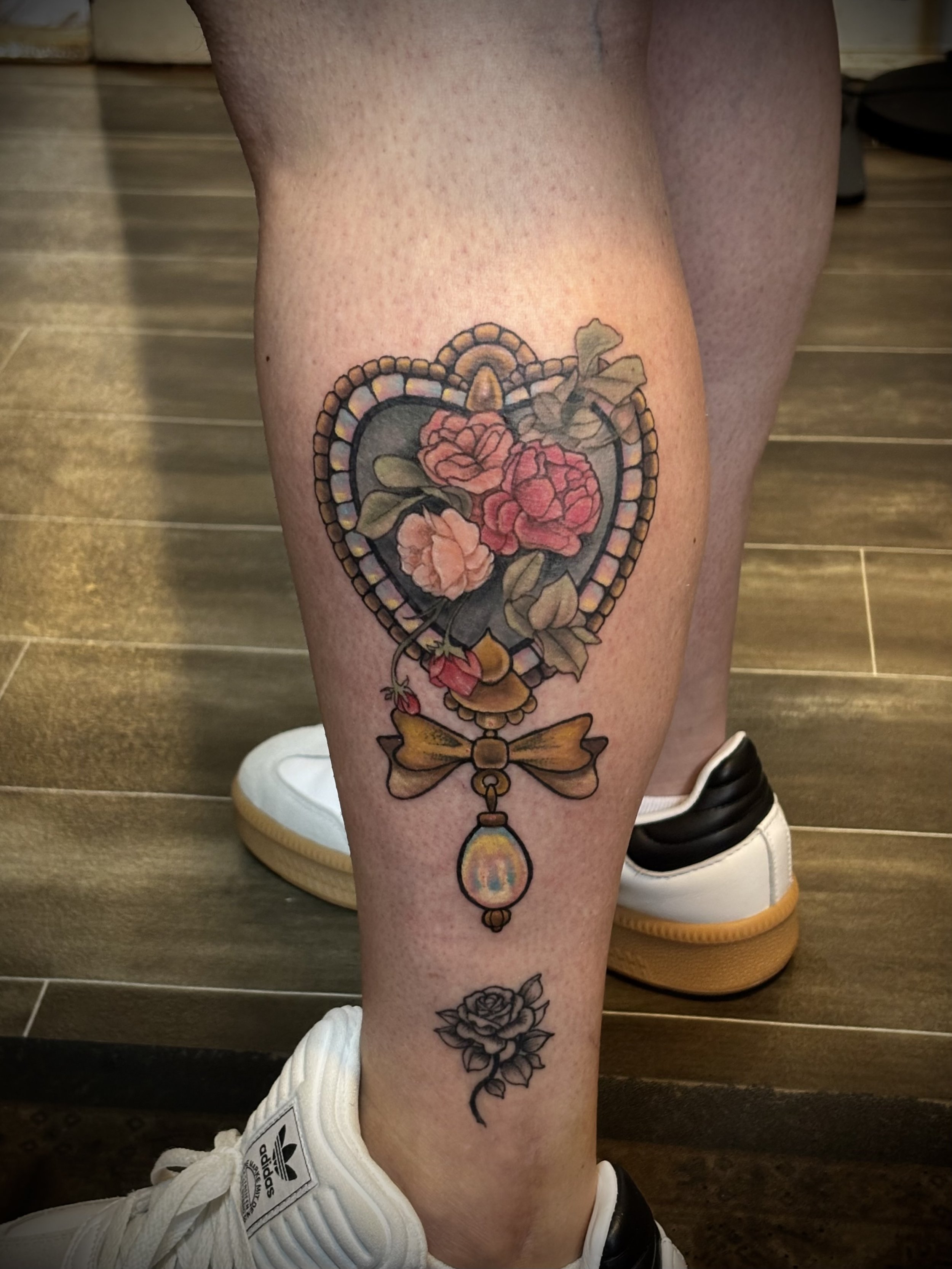 Tattoo of an ornamental gold heart with iridescent inlays, opal gem charm and baroque flowers on a leg