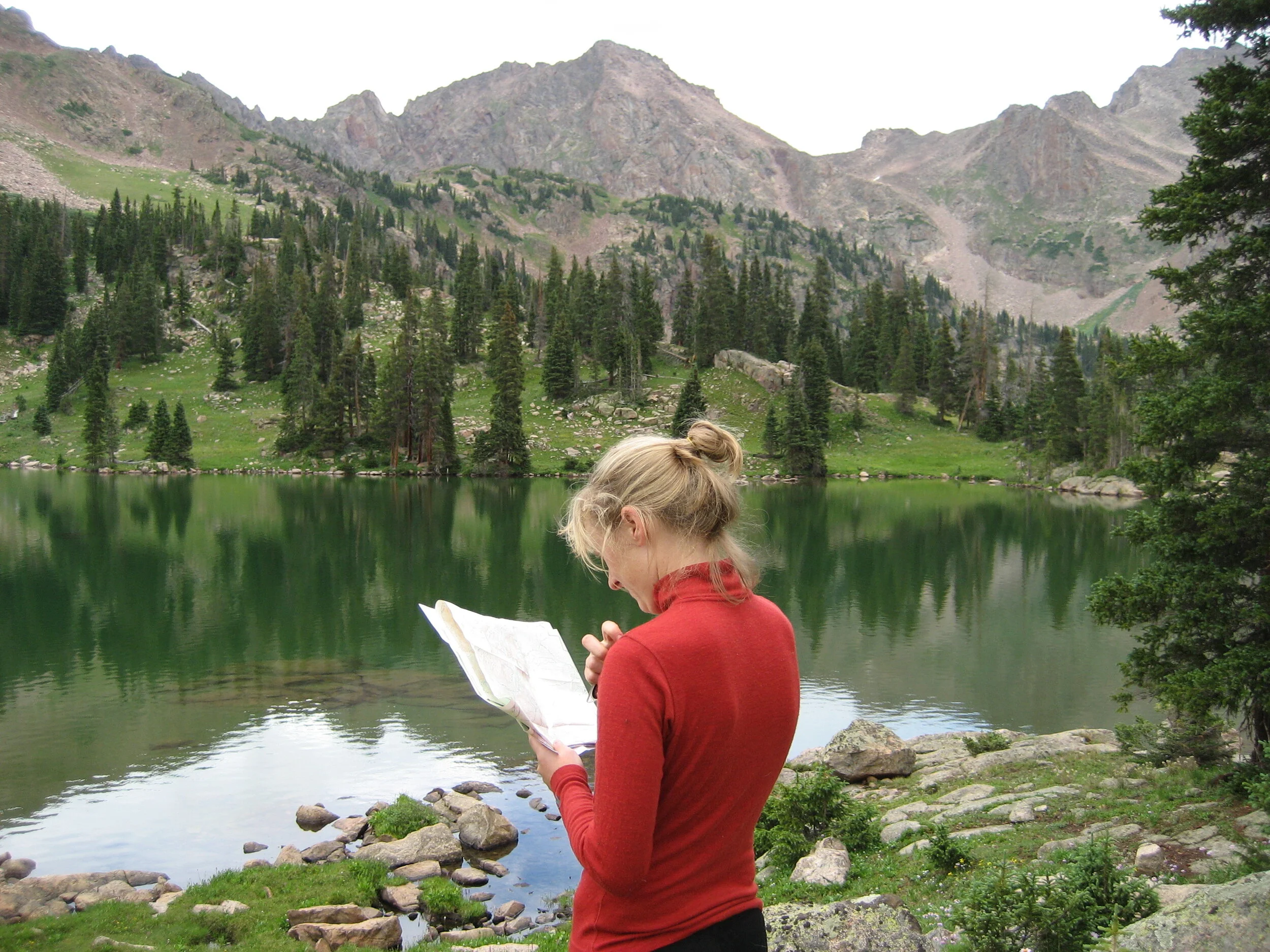 A person with blonde hair tied up in a bun, wearing a red long-sleeved shirt, stands near a body of water reading a map, with lush green trees, rocky shore, and tall mountain peaks in the background.